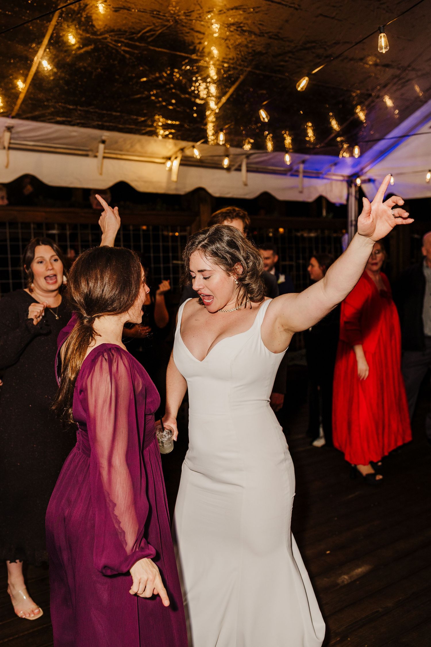 Bride in white gown dances energetically with guests at indoor wedding reception with string lights.