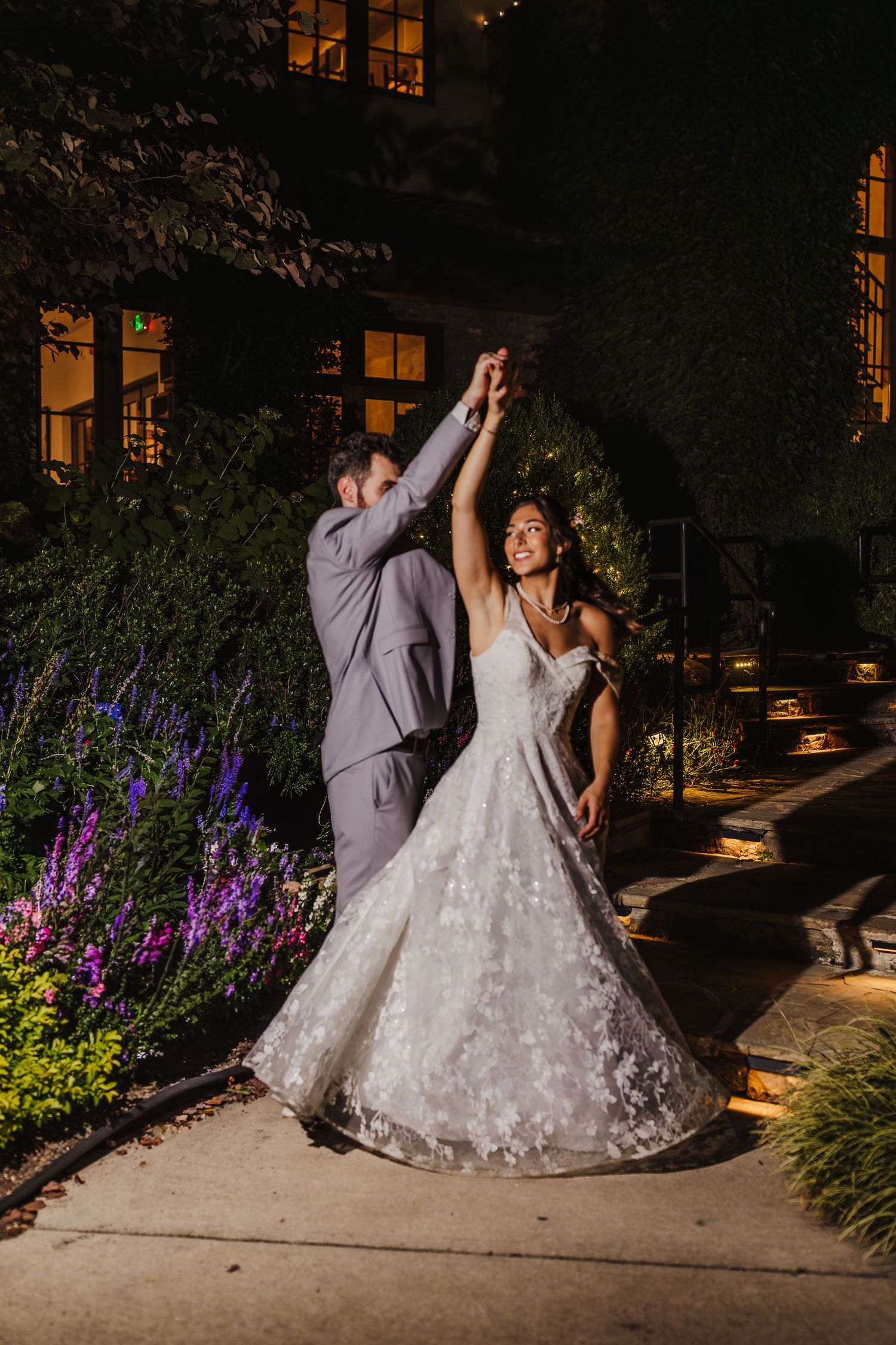 Bride and groom dance outdoors at night, groom twirling bride in a floral lace gown near a lit building.