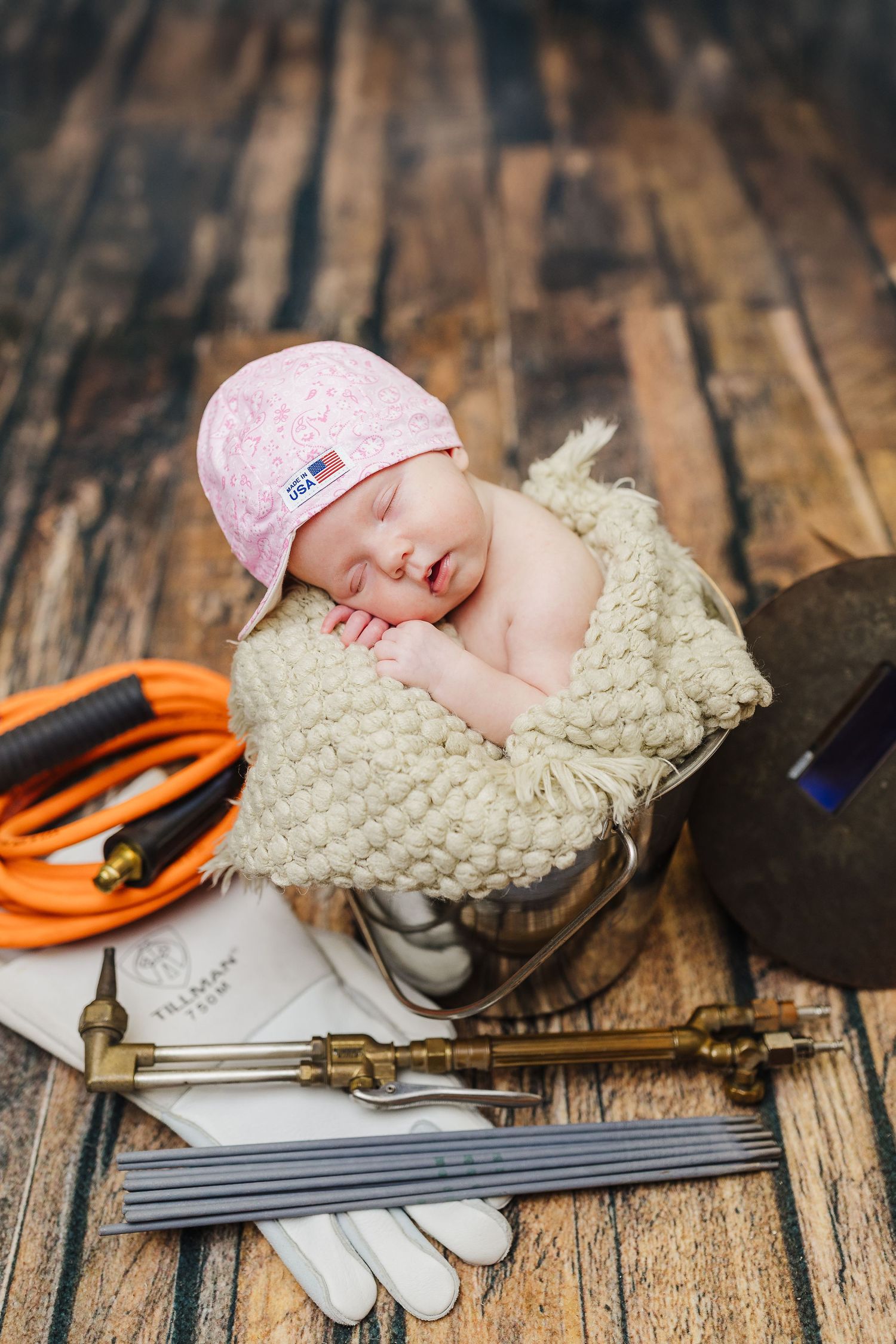 Sleeping baby in pink hat nestled in cream blanket with baseball equipment on rustic wooden surface.