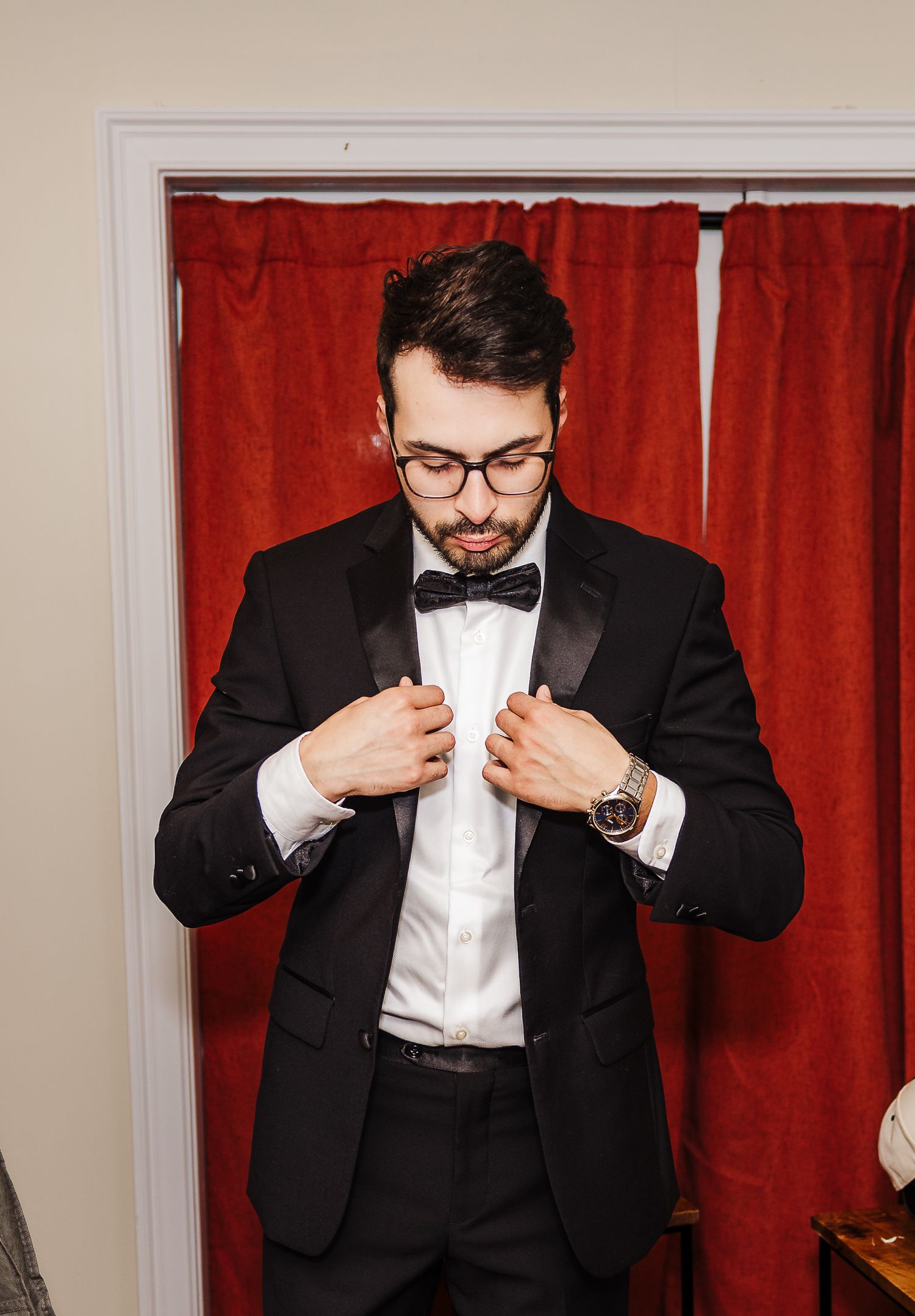 Person in formal black tuxedo adjusts bowtie while standing against red curtain.