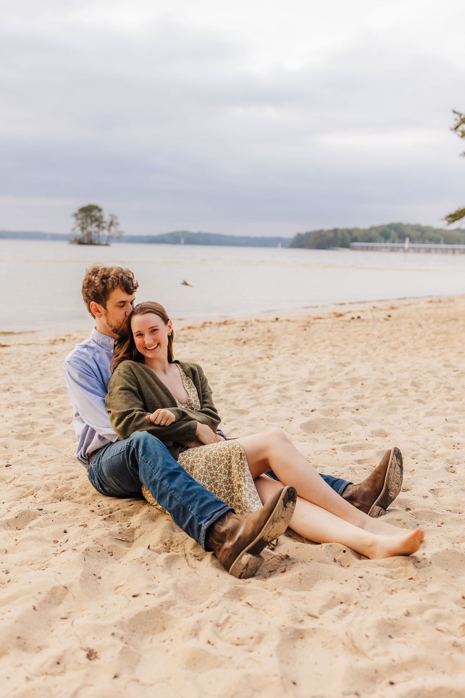 Couple relaxing together on sandy lake shore with water and trees in background.