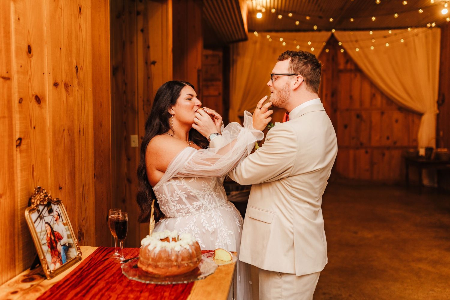 Bride and groom feeding each other wedding cake in a rustic barn venue with warm string lights.