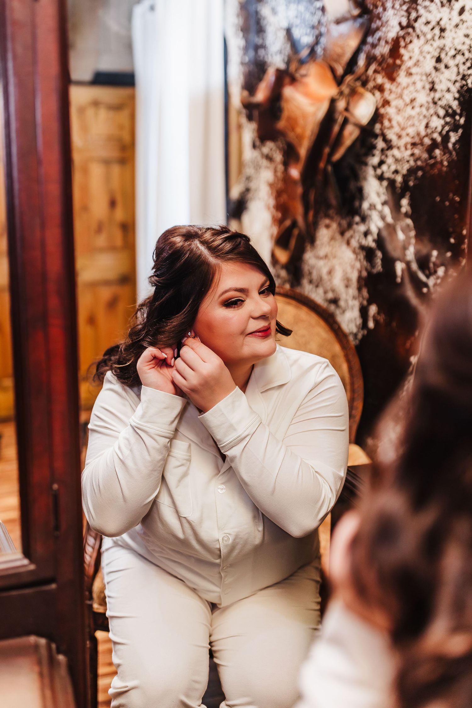 Bride in white suit adjusting earring while seated, reflected in mirror during wedding preparation.