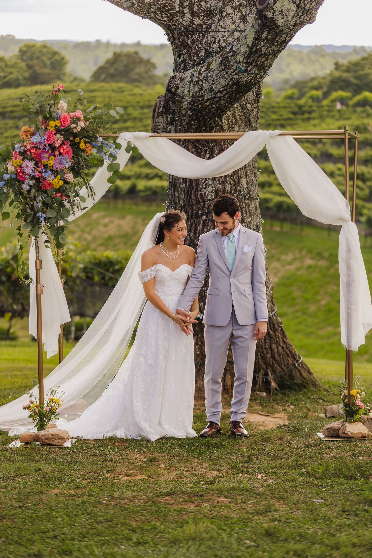Bride and groom stand under floral arch with white draping beside large tree at outdoor vineyard wedding ceremony.