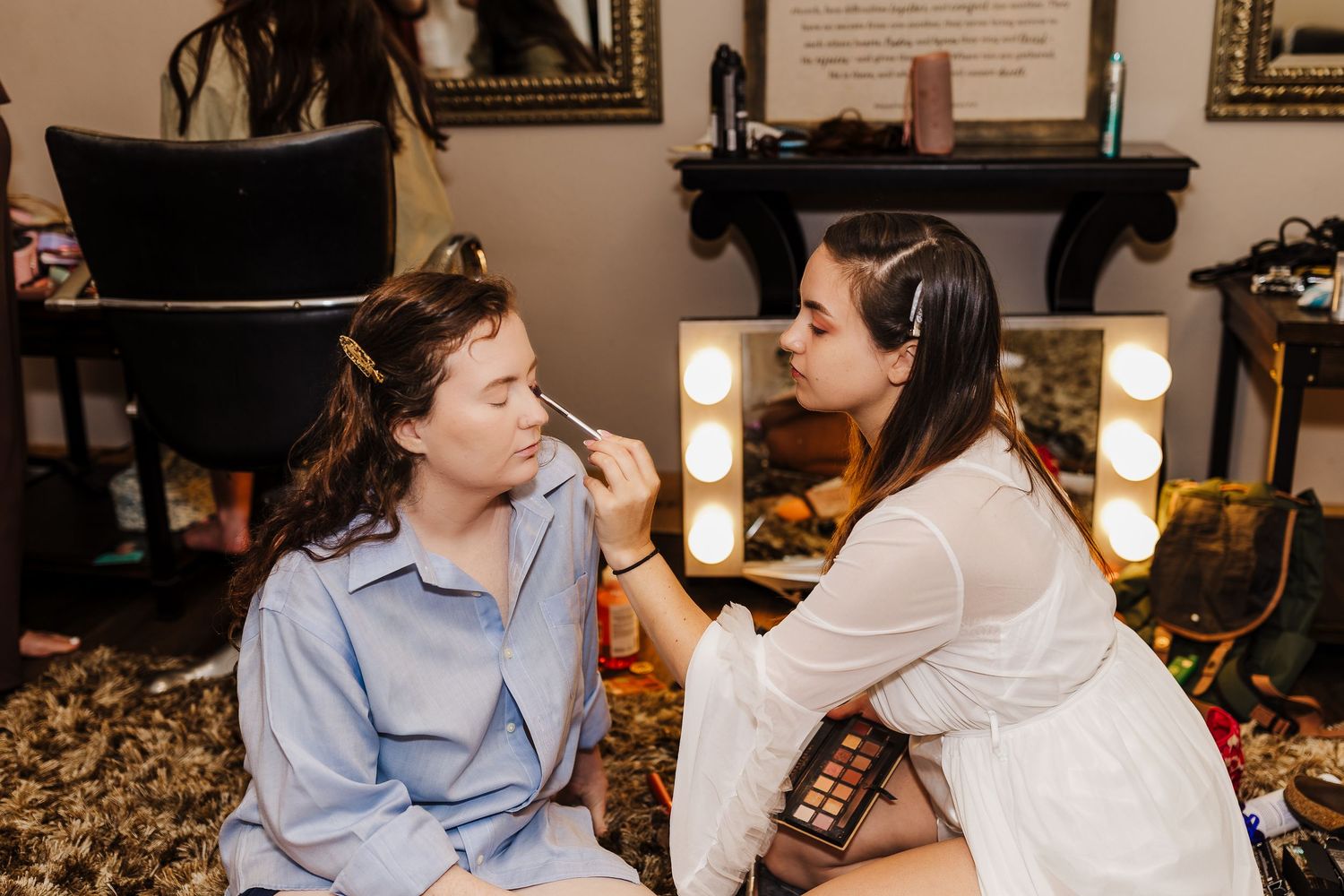 A makeup artist applies cosmetics to a client in a dressing room with illuminated vanity mirrors.