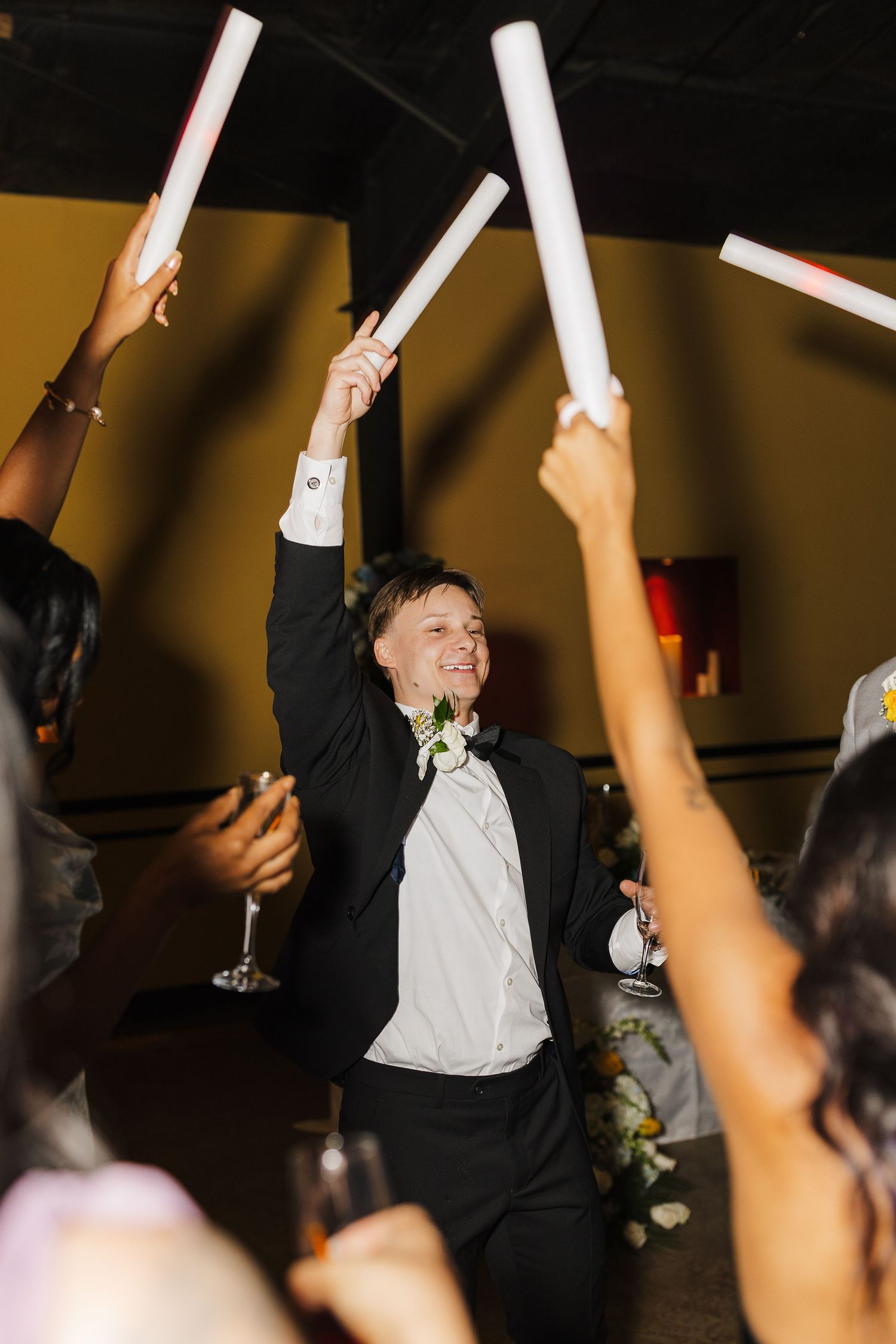 Groom in black tuxedo celebrates with guests holding white glow sticks at a wedding reception.
