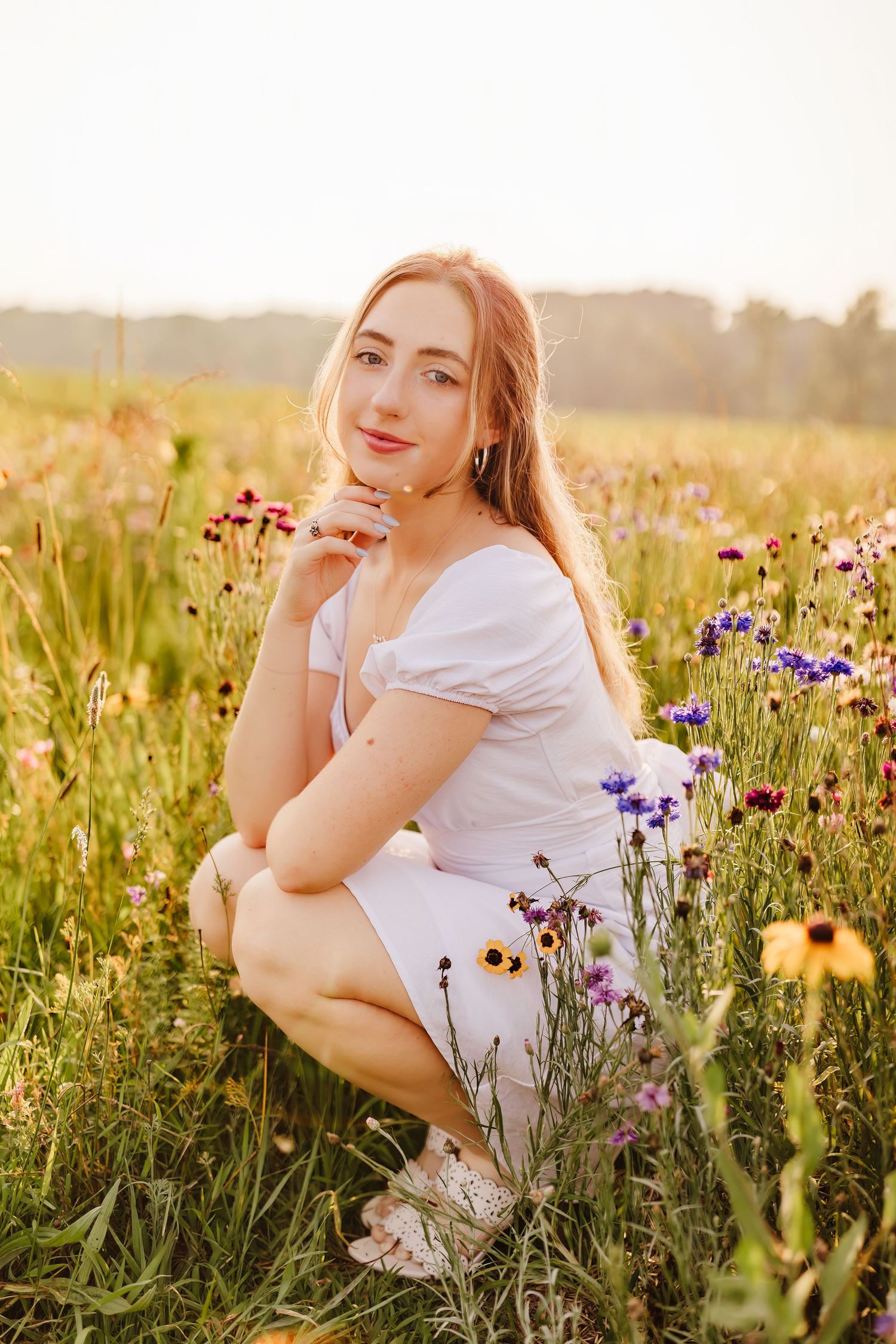 Young woman in white dress crouching in a wildflower meadow at golden hour sunset.