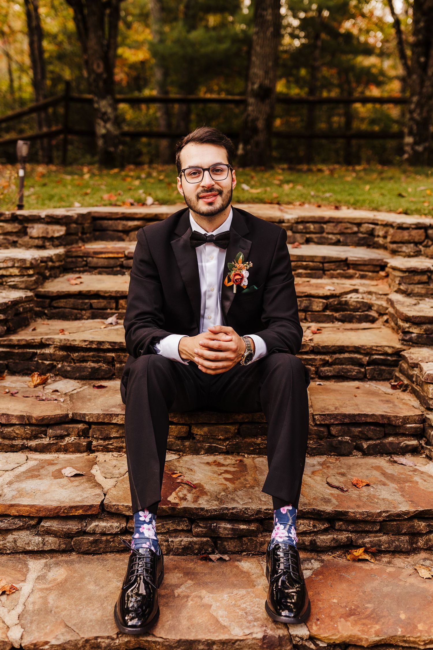 Groom in black suit and bow tie sits on rustic stone steps wearing unique patterned socks.