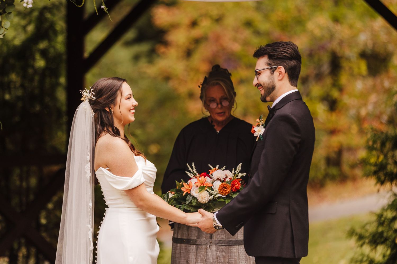 Bride and groom hold hands during outdoor wedding ceremony with officiant and colorful autumn foliage backdrop.