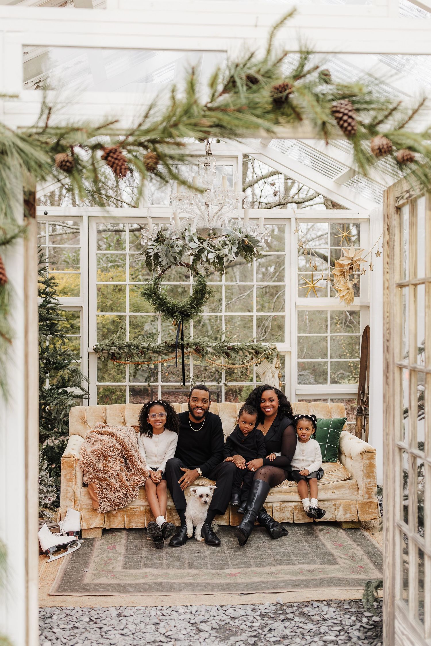 Family of four with a dog poses on a vintage sofa in a festive greenhouse decorated for Christmas.