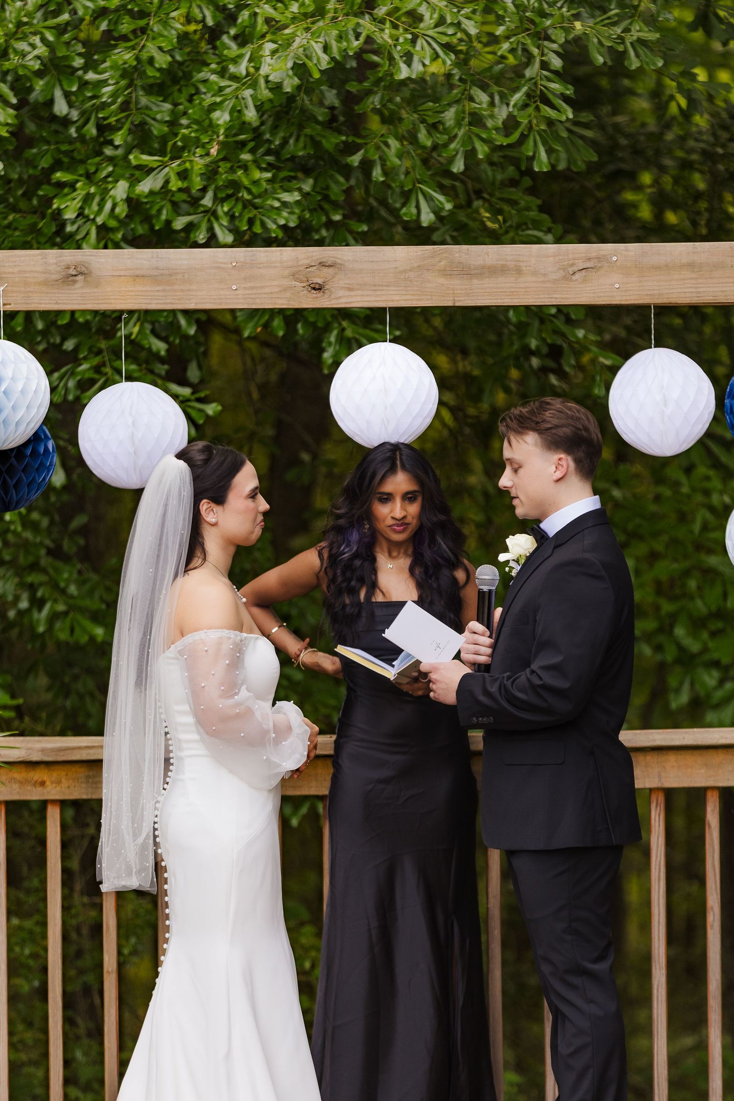 Groom reads vows to bride in white gown and veil as officiant stands between them under decorated wooden arch outdoors.