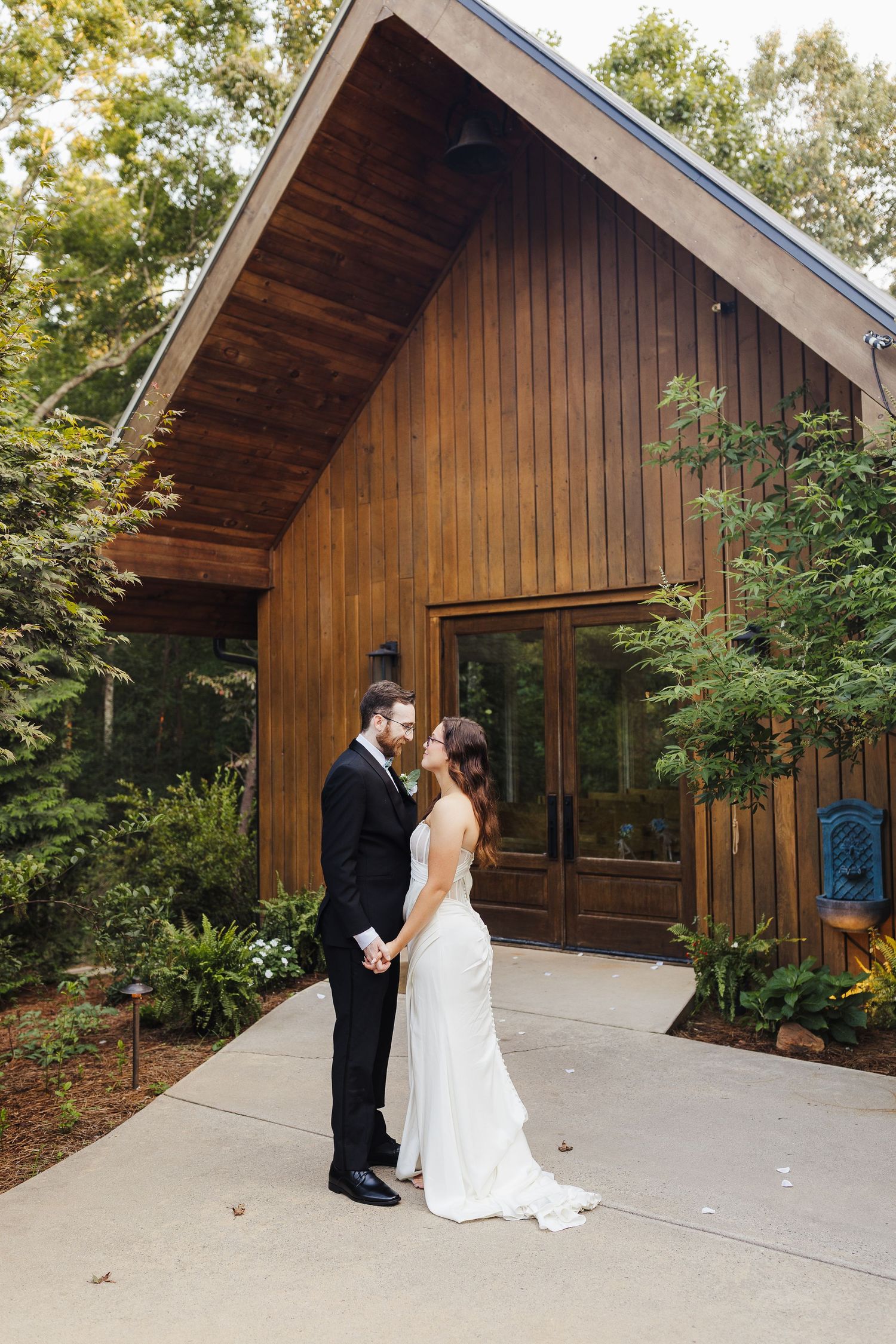 Bride and groom hold hands facing each other outside a modern wood cabin wedding venue surrounded by lush greenery.