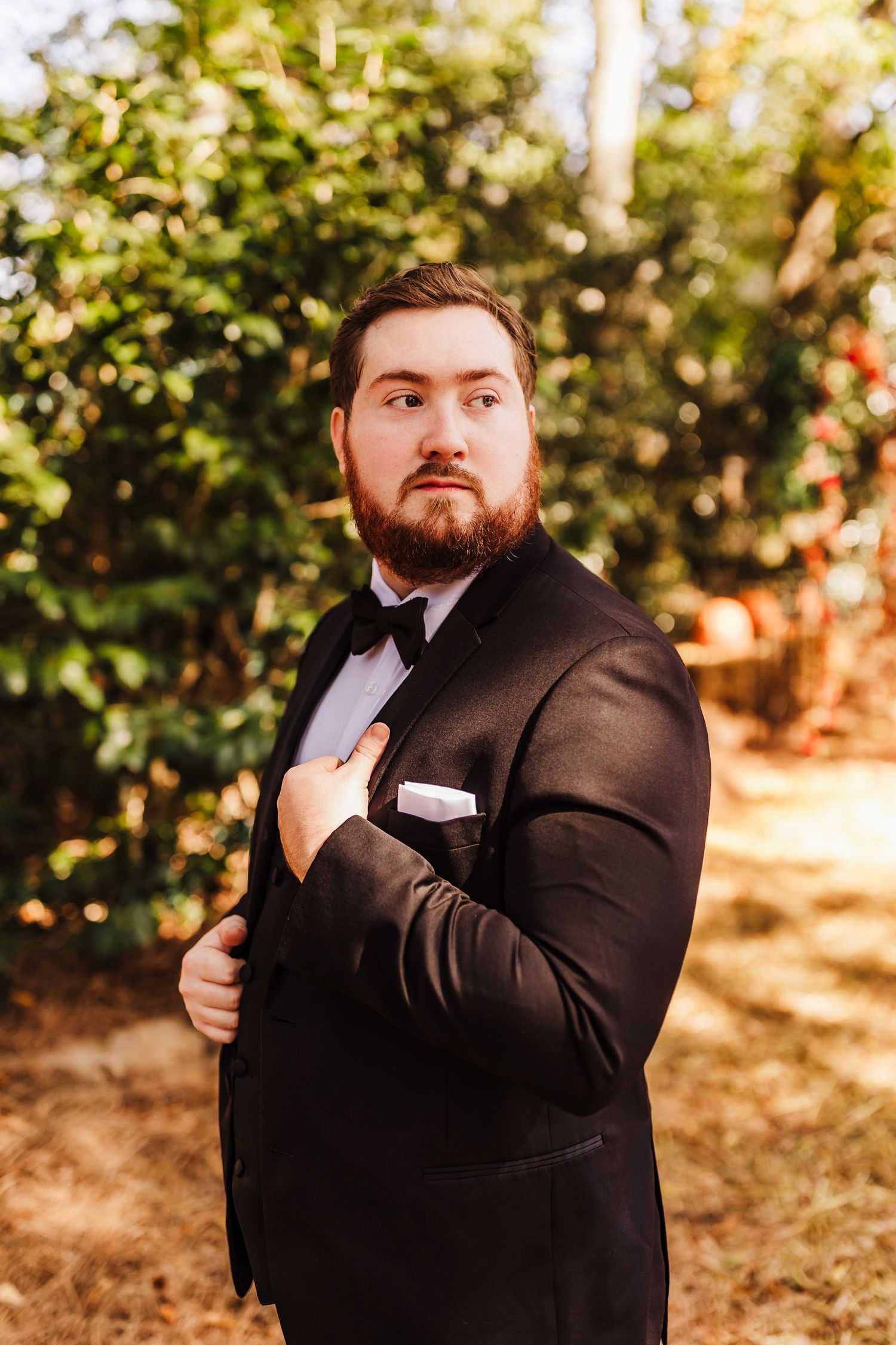 Groom in classic black tuxedo poses outdoors in autumn setting with natural bokeh background.