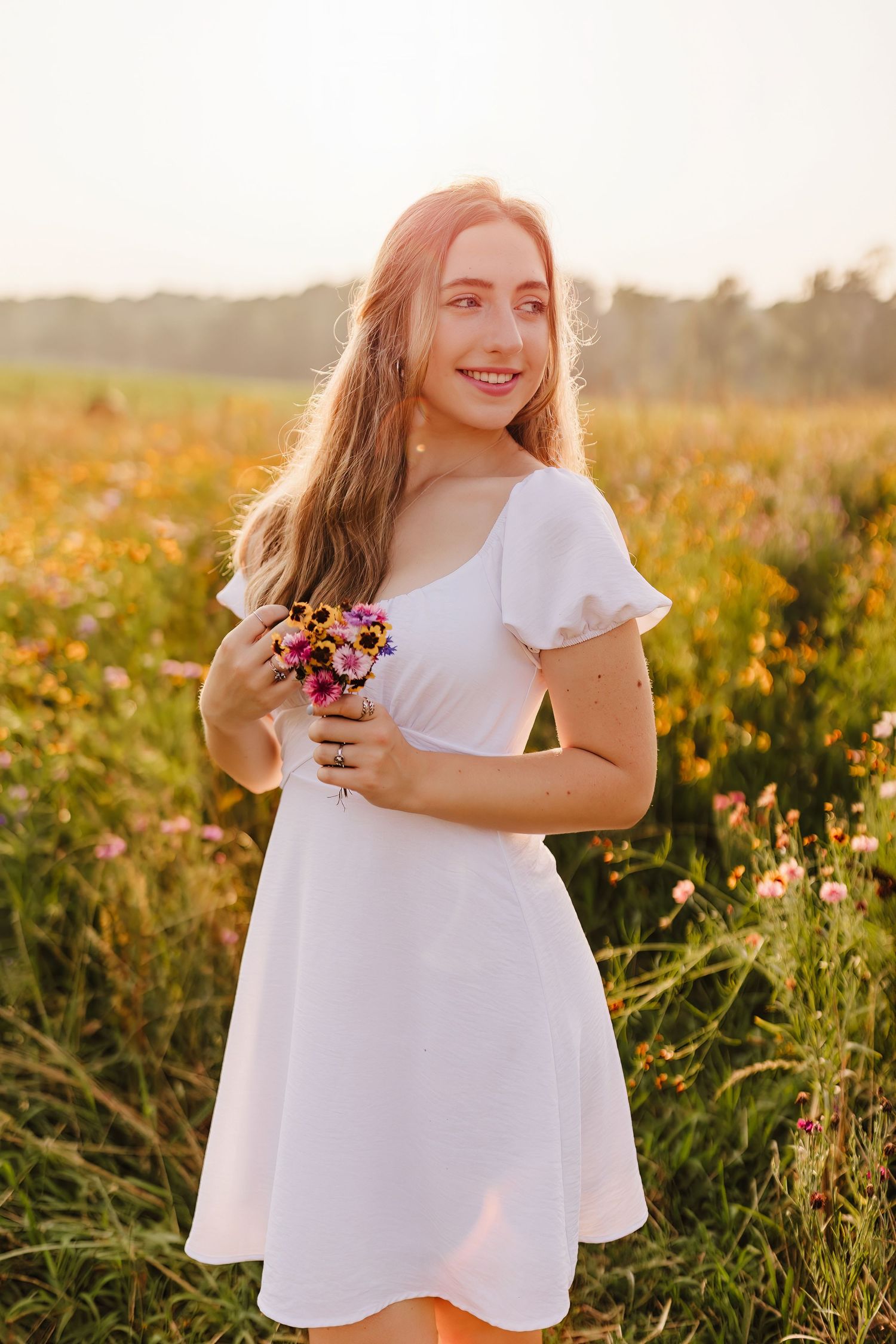 Young woman in white dress holding wildflowers smiling in a sunlit meadow of colorful blooms at golden hour.