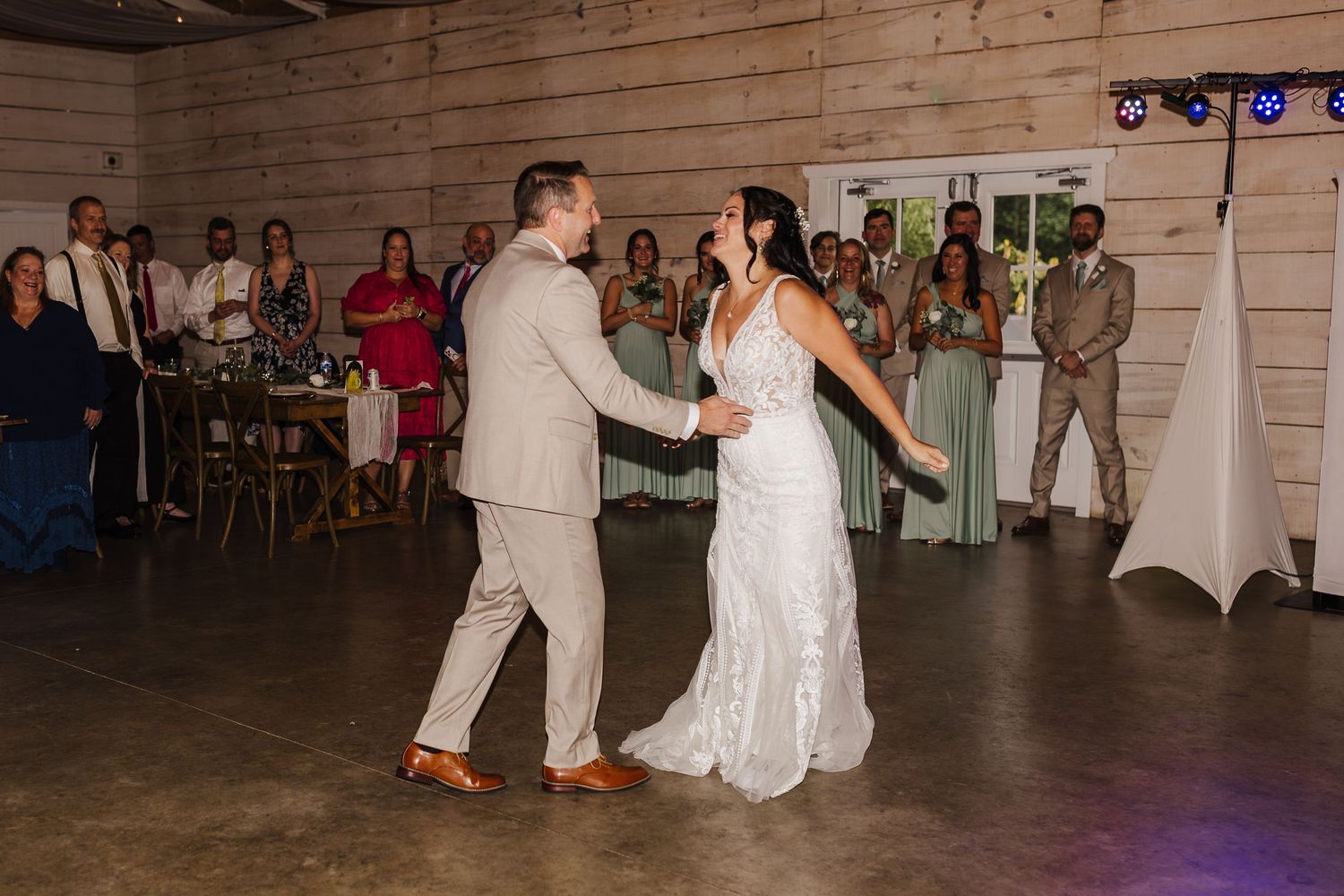 Bride and groom share their first dance at a rustic wedding reception with guests applauding in the background.