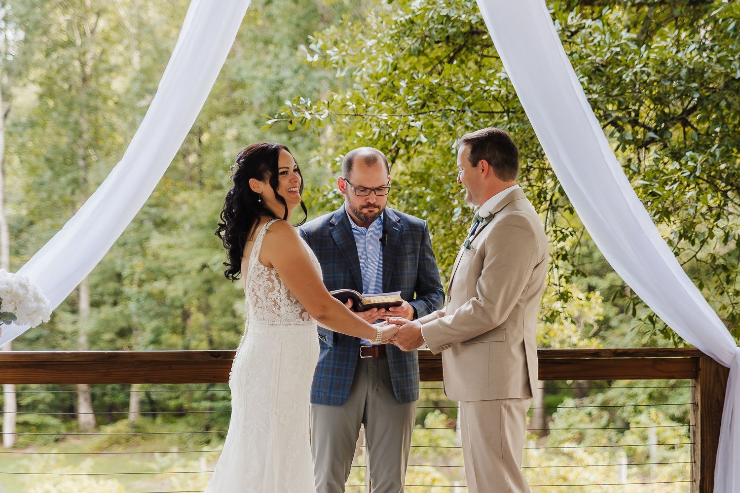 Outdoor wedding ceremony with bride and groom exchanging vows before officiant under white draped arch surrounded by trees.