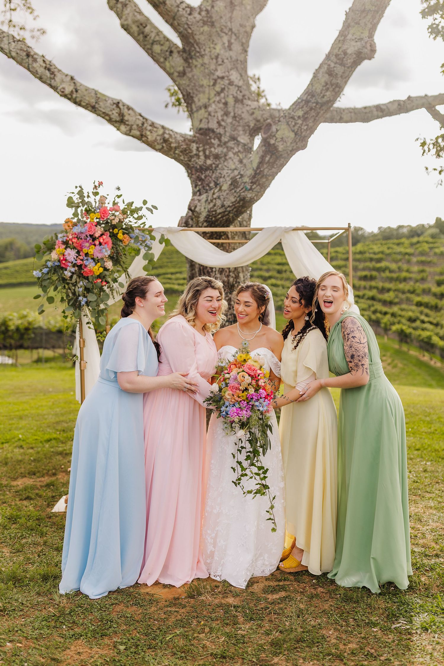 Bride and four bridesmaids in pastel dresses laughing outdoors at vineyard wedding ceremony under decorated arch.