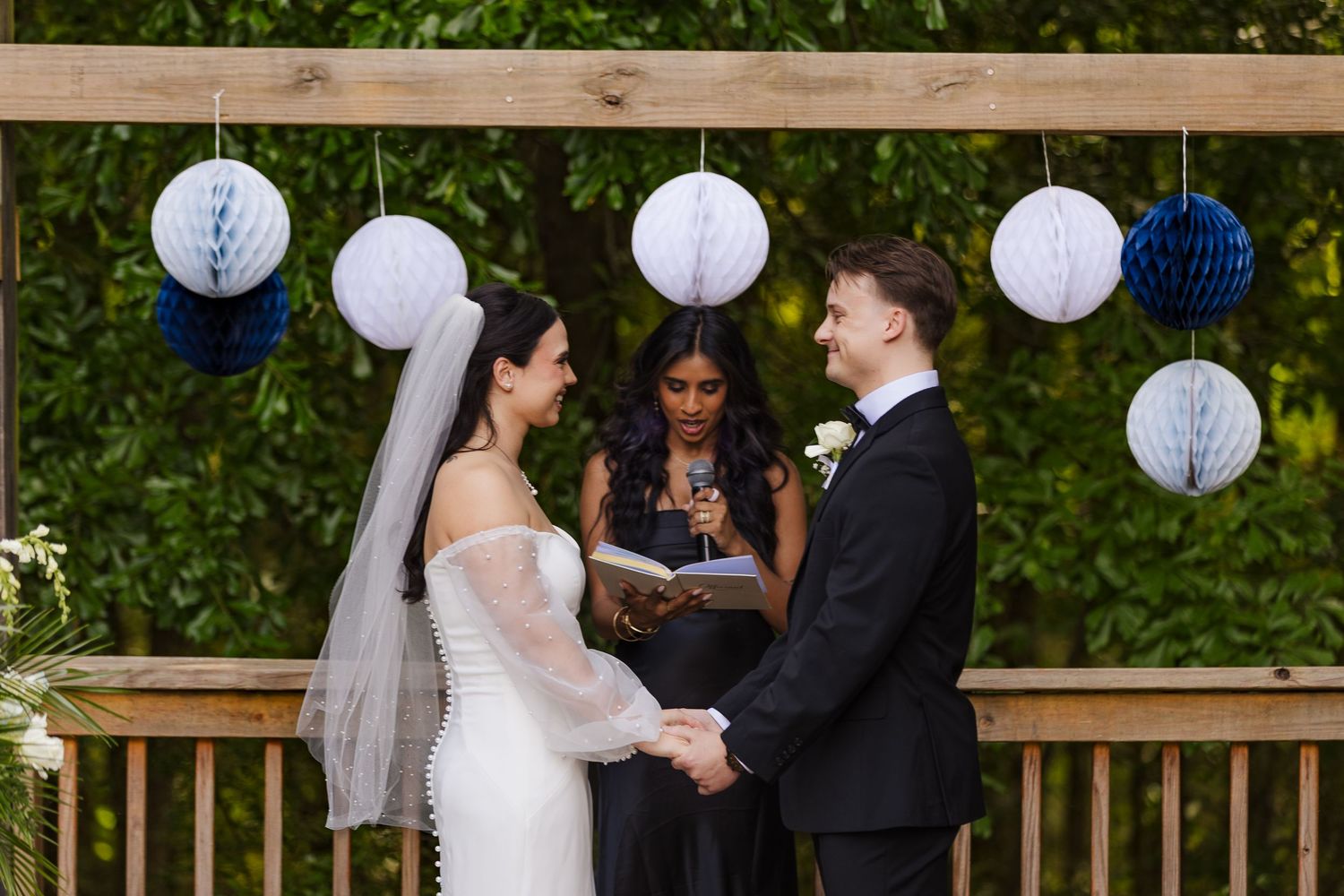 Bride and groom exchange vows outdoors under wooden arch with blue and white honeycomb paper lanterns, officiant reading ceremony script.