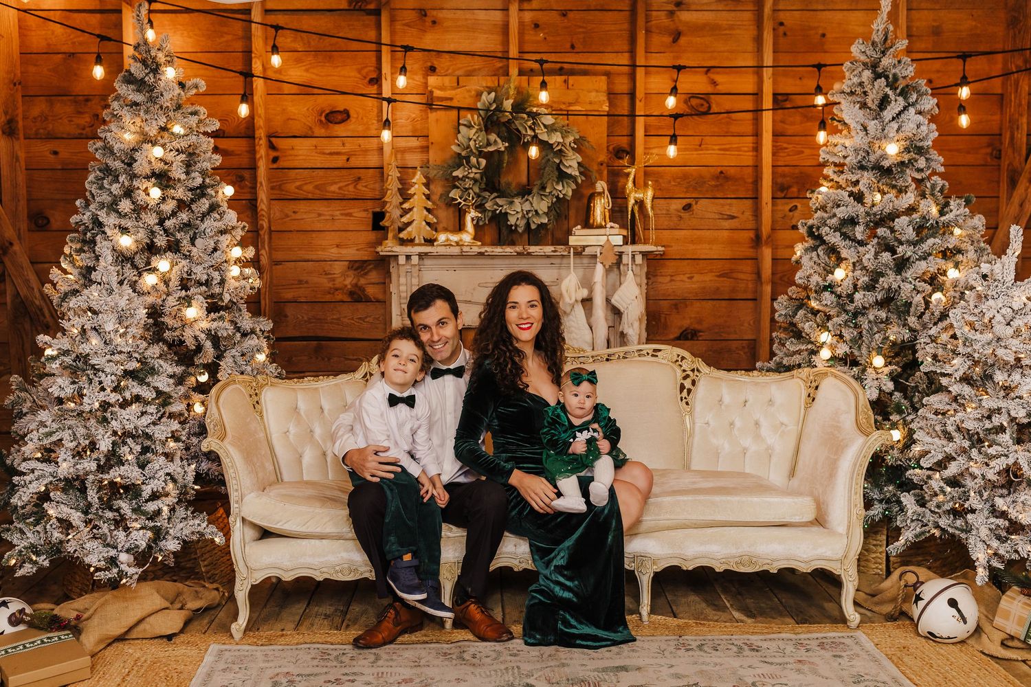 Family of four poses on white sofa between flocked Christmas trees in rustic wood-paneled holiday photo studio.