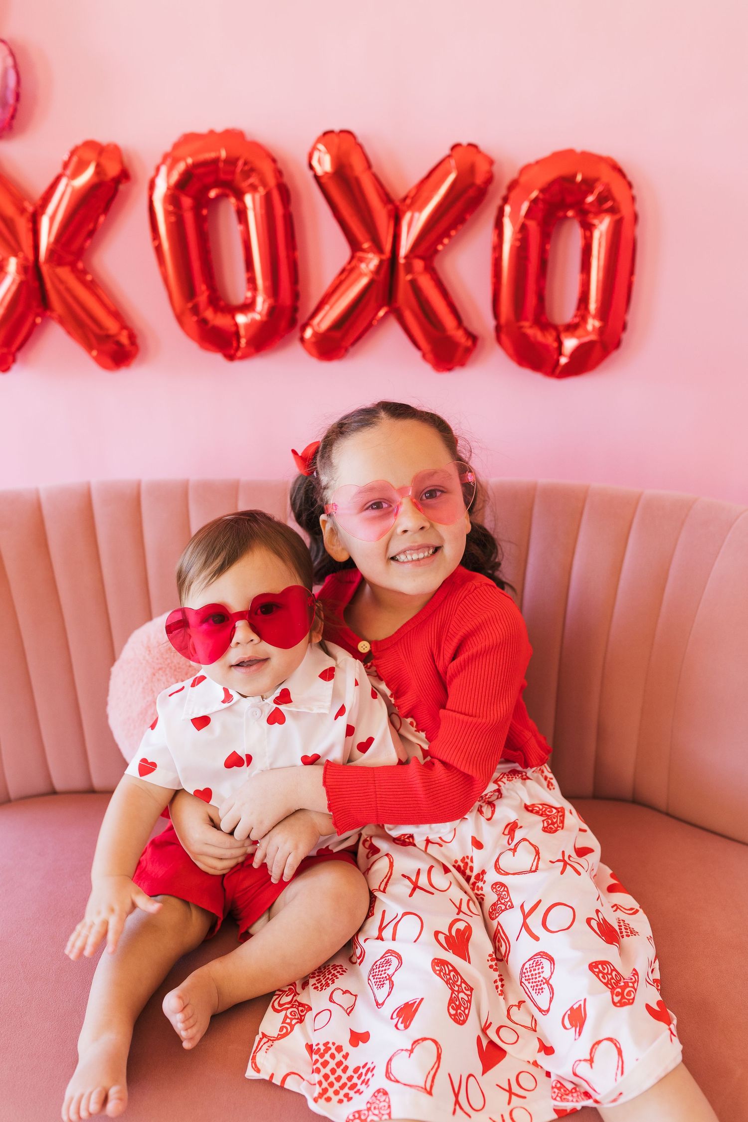 Two young girls in Valentine's Day outfits pose together under red XOXO balloons on a pink couch.