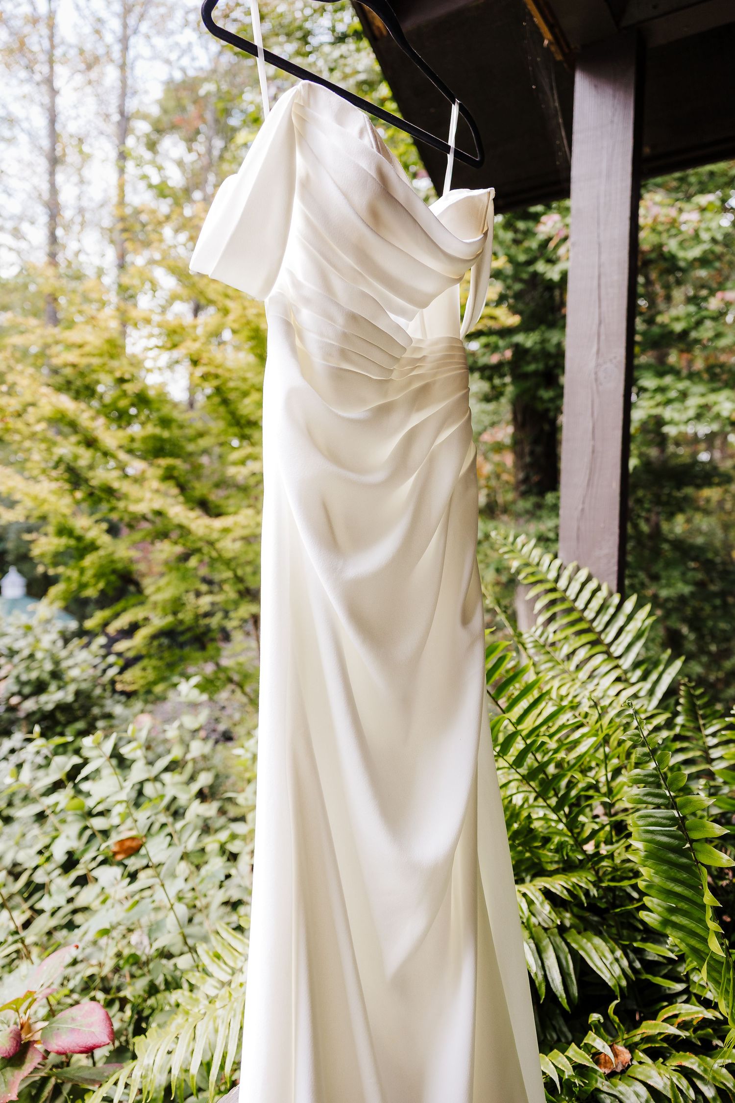 White wedding dress hangs elegantly against lush green ferns and garden foliage in outdoor setting.