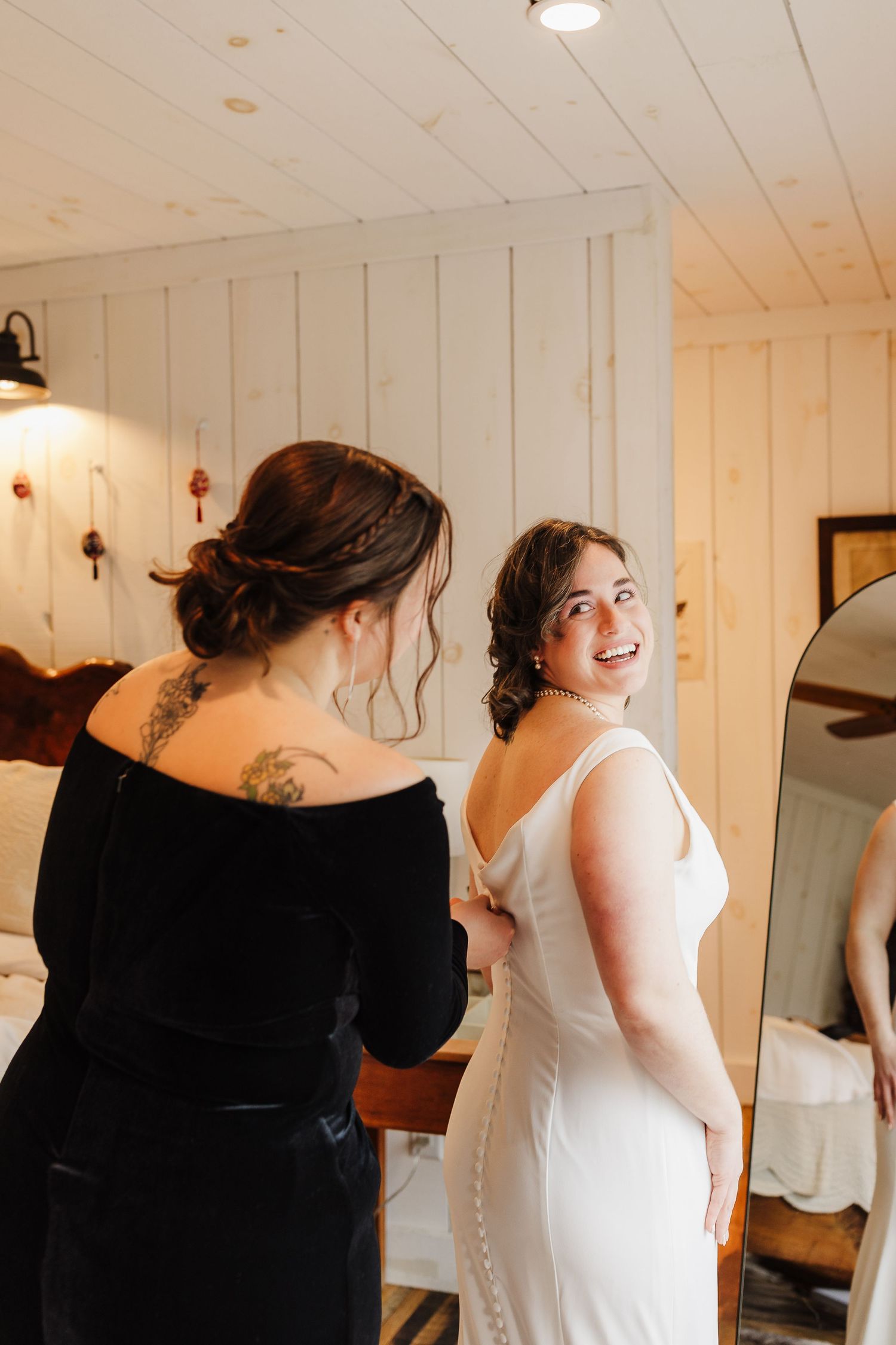 Bride smiling over shoulder as bridesmaid buttons up white wedding gown in rustic white wood-paneled room.