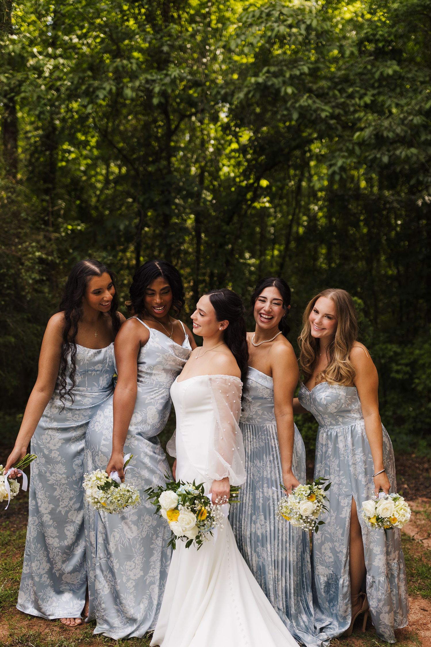 Bride laughing with four bridesmaids in blue floral gowns holding white bouquets outdoors in a lush green forest setting.