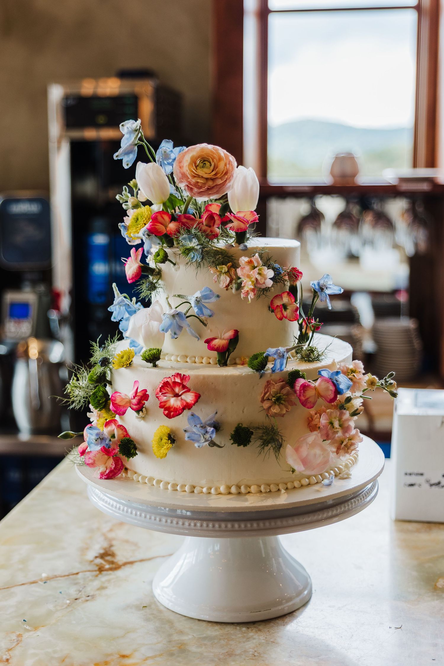 Elegant three-tier white wedding cake adorned with colorful fresh flowers including roses and blue blooms on marble stand.