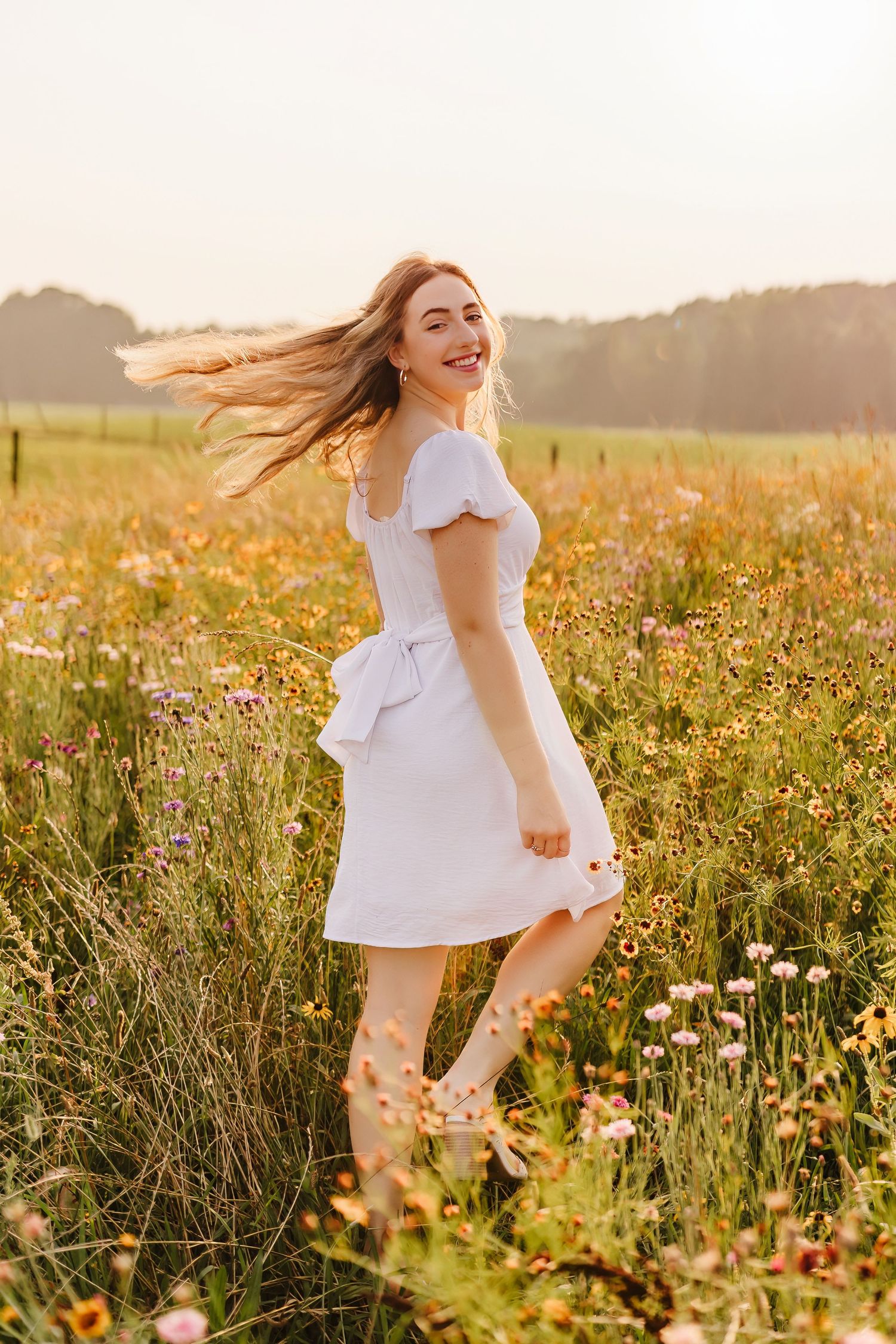 Young woman in white dress laughing in a wildflower meadow at golden hour, hair blowing in the breeze.