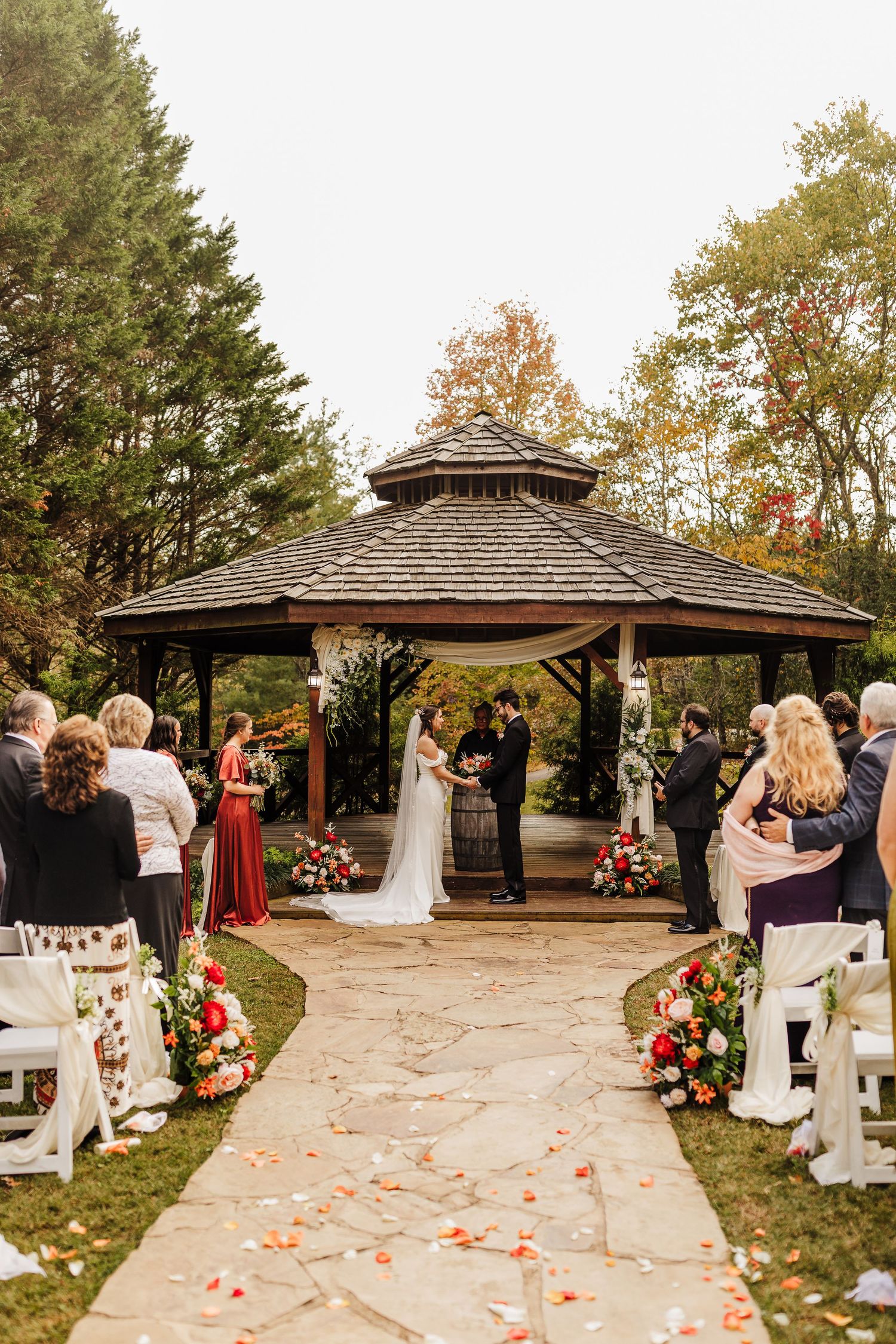Outdoor wedding ceremony under a wooden gazebo surrounded by fall foliage and scattered rose petals on stone path.