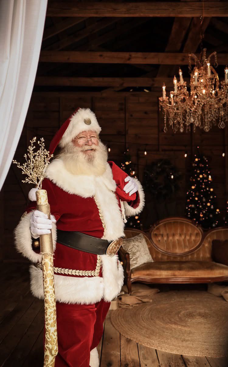 Santa Claus in red velvet suit holding golden staff in rustic wooden cabin with chandelier and Christmas tree.