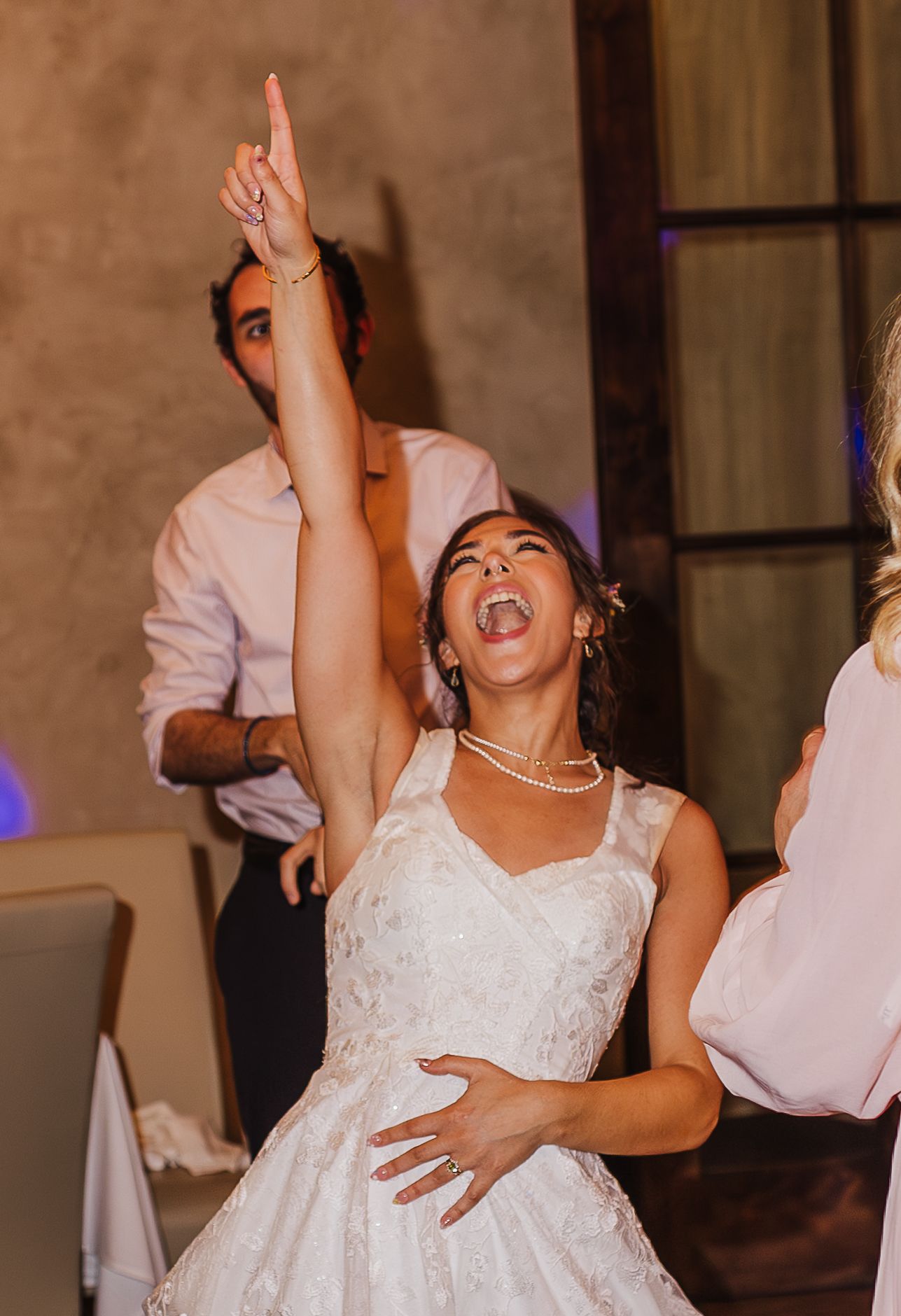 Bride in white lace dress dancing joyfully with arm raised at wedding reception.