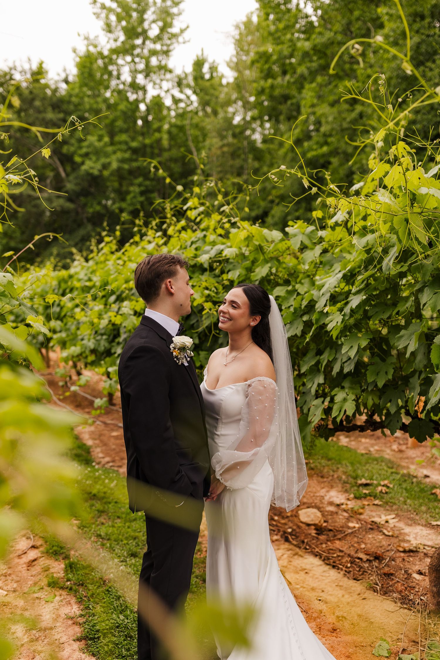 A bride and groom share a romantic moment among lush green vineyard rows, the bride smiling in a white off-shoulder gown and veil.