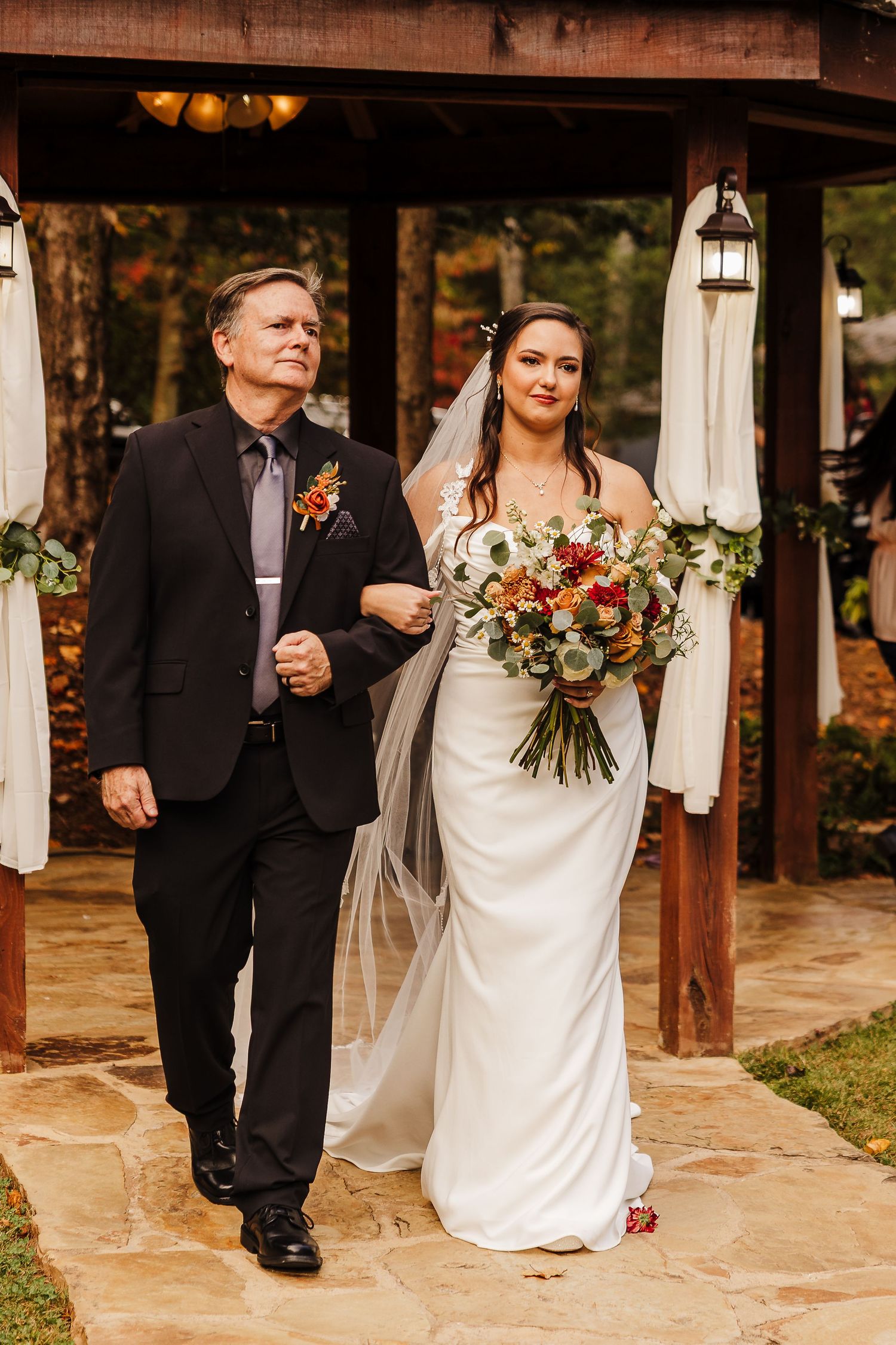 Wedding party walks along a stone path decorated with white drapes and lanterns at an outdoor venue.