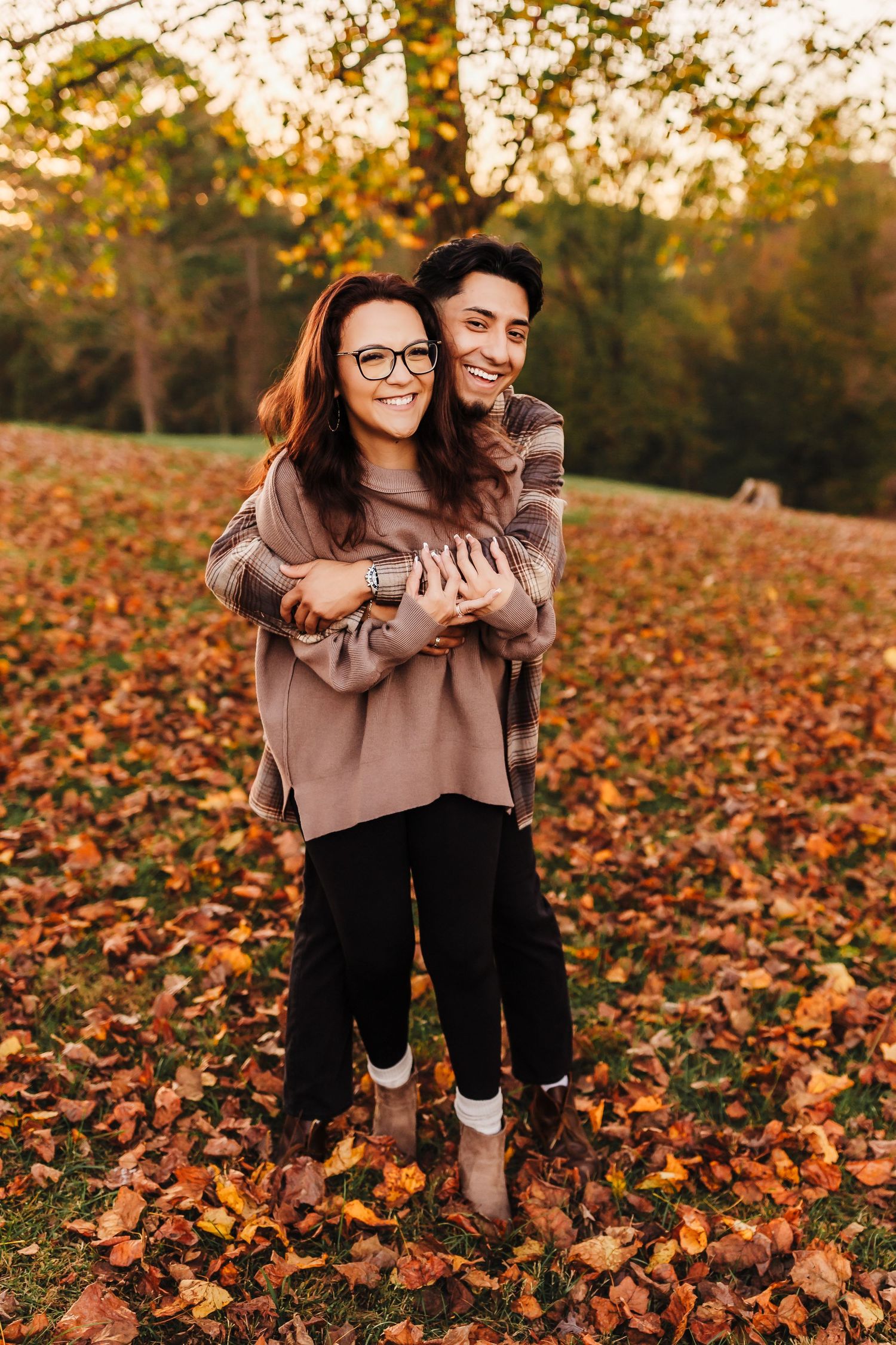Couple embracing among fallen autumn leaves in warm sweaters during sunset photoshoot.
