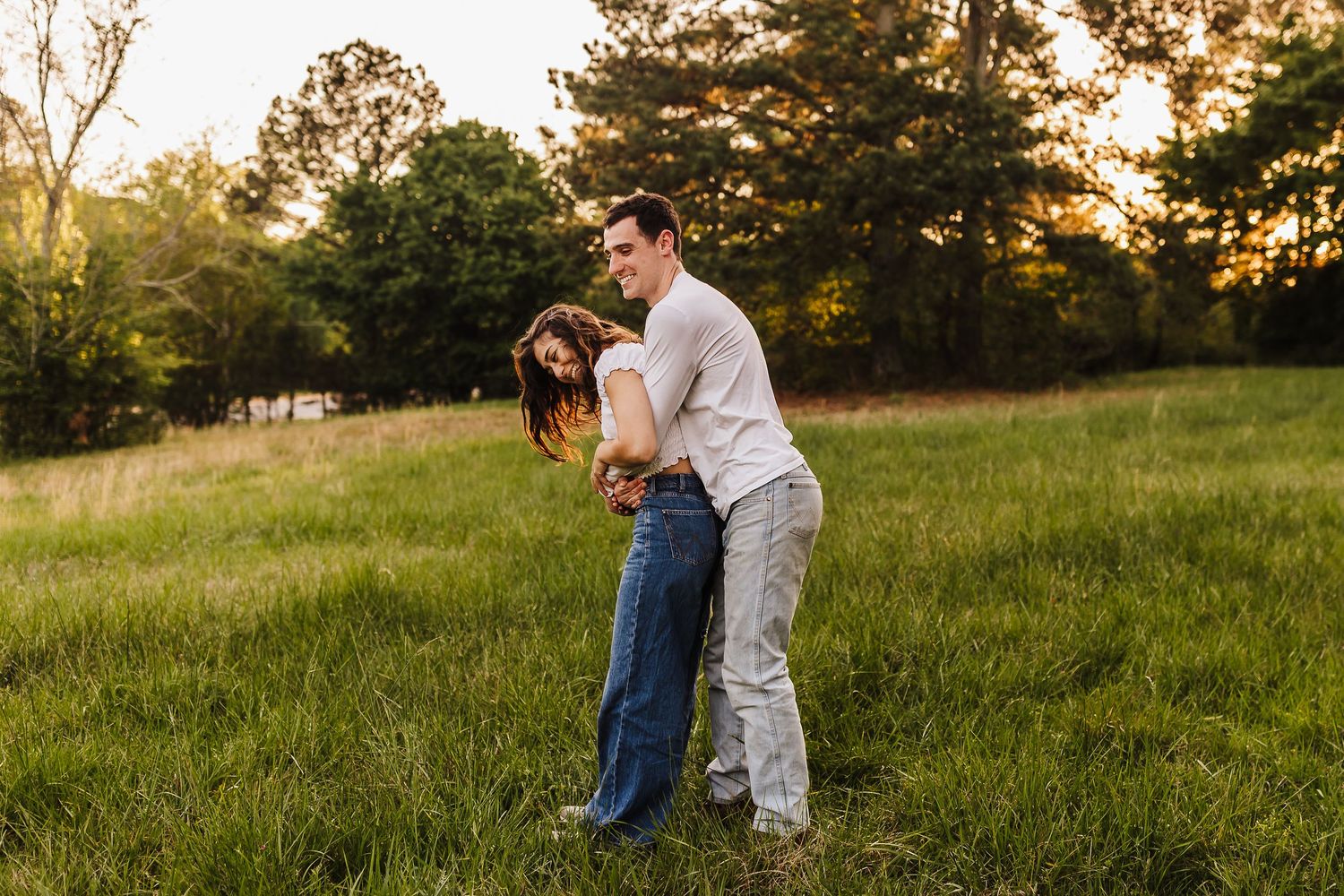 Couple embraces in a sunlit meadow during golden hour as warm light filters through trees in the background.