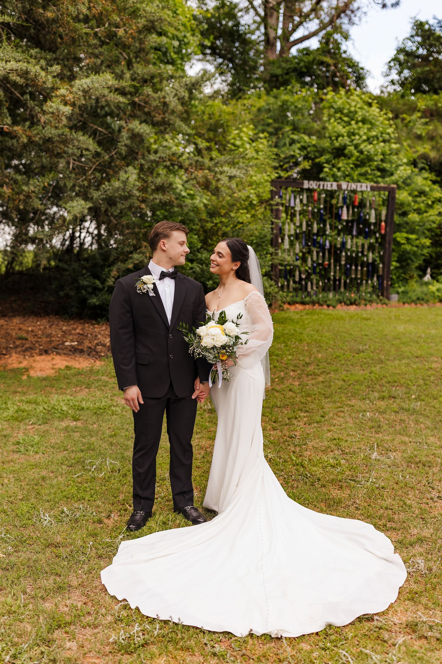 Bride and groom holding hands in white gown and black tuxedo outdoors at a winery wedding venue surrounded by lush trees.