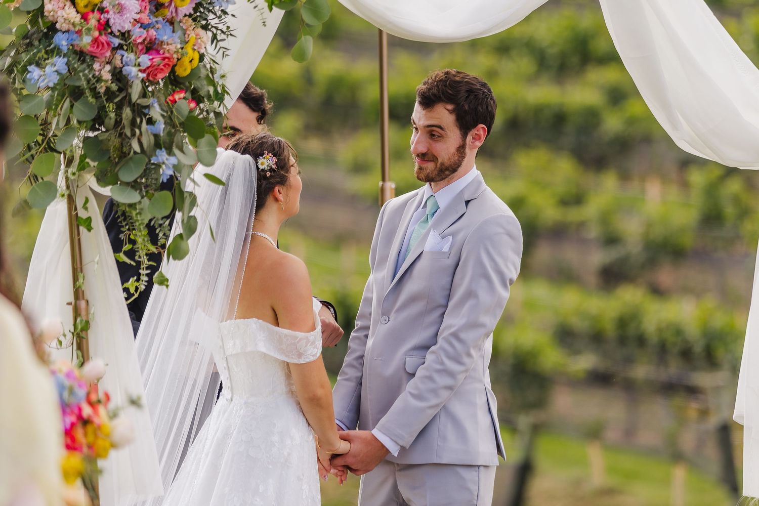 Bride and groom holding hands at outdoor vineyard wedding ceremony beneath floral arch with colorful blooms.