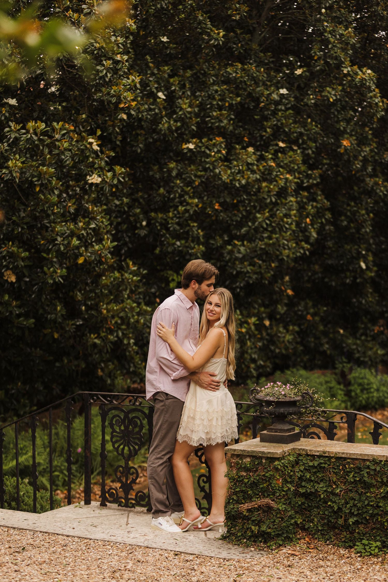 Couple embracing tenderly in an elegant garden with ornate iron railing and dense magnolia trees.