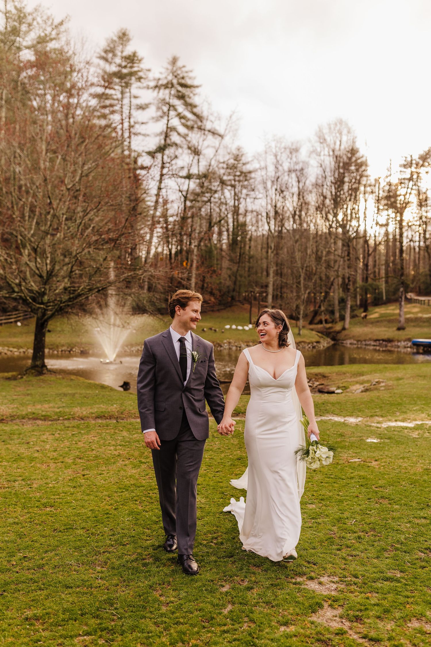 Bride and groom walk hand-in-hand on green lawn with pond fountain and trees in background.
