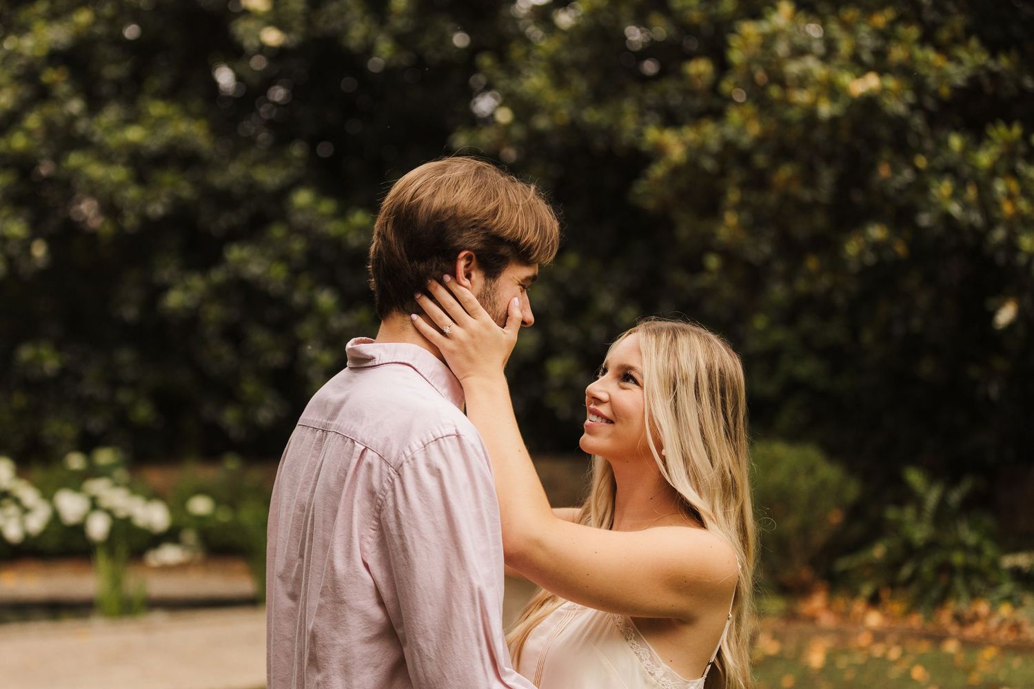 A couple shares a romantic moment outdoors, with the woman tenderly holding the mans face while smiling at each other.