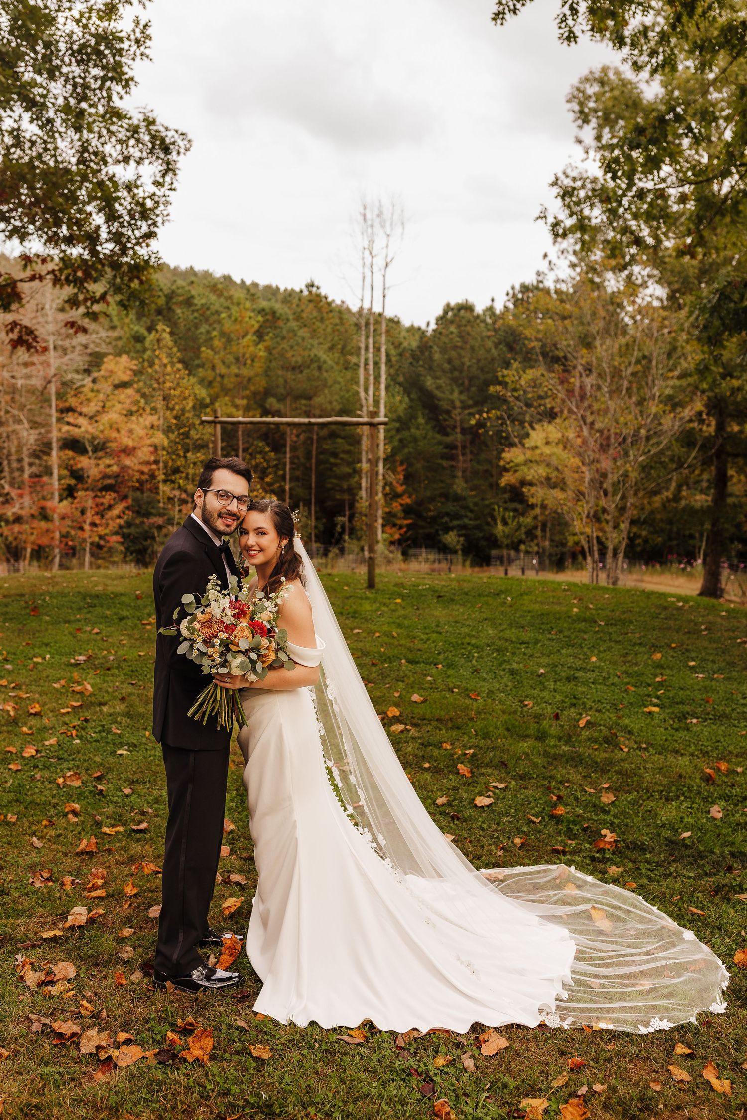 Bride and groom pose in autumn meadow, her cathedral veil trailing on fallen leaves, with colorful fall foliage backdrop.