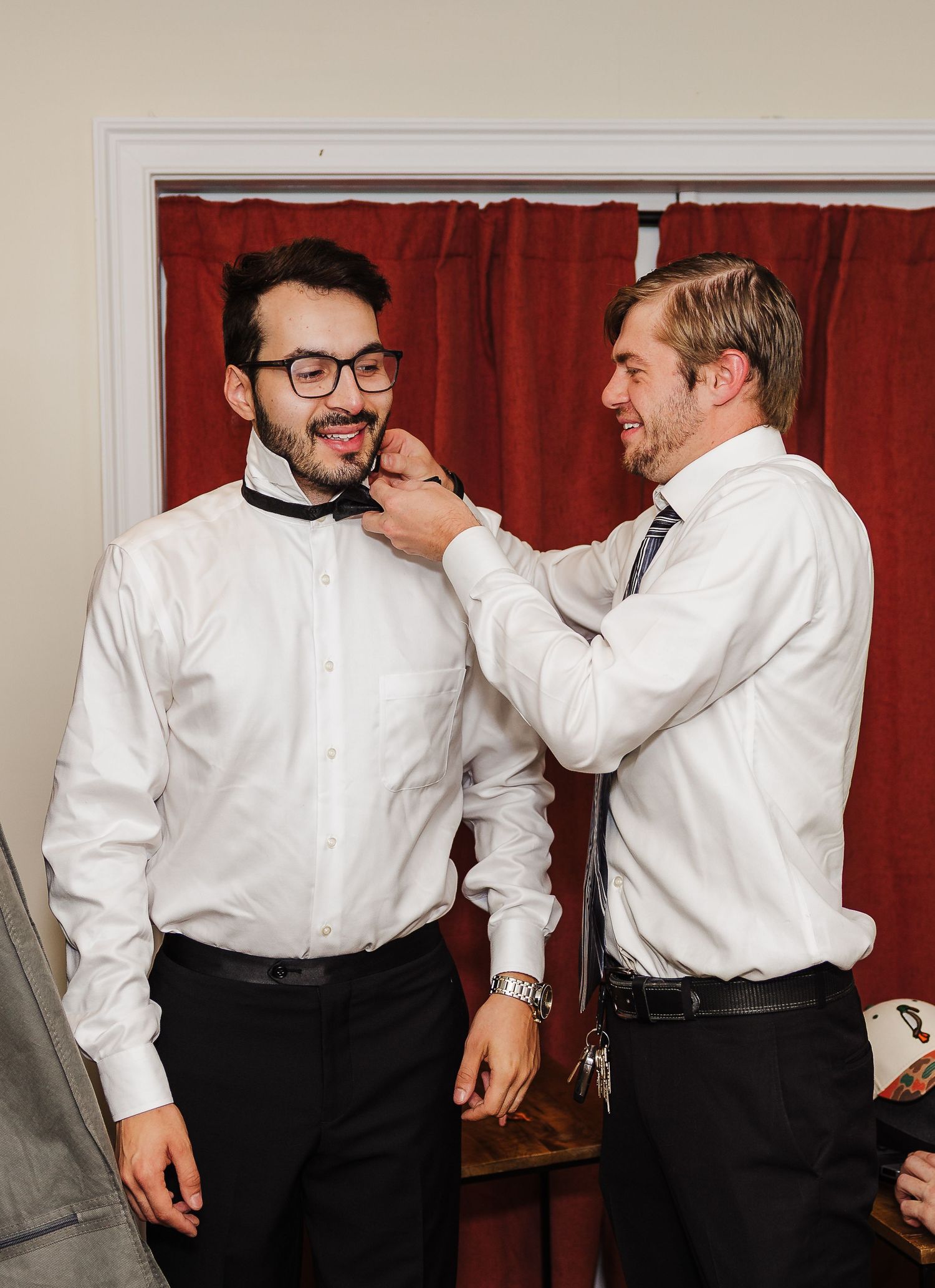 Two people in white dress shirts share a playful moment in front of a red curtain backdrop.