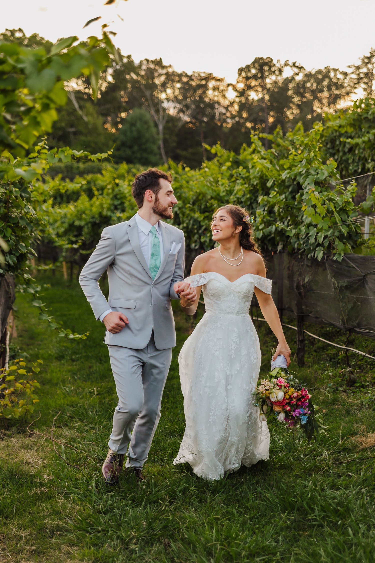 A smiling bride and groom walk hand-in-hand through a lush vineyard, she in a white gown holding a colorful bouquet.