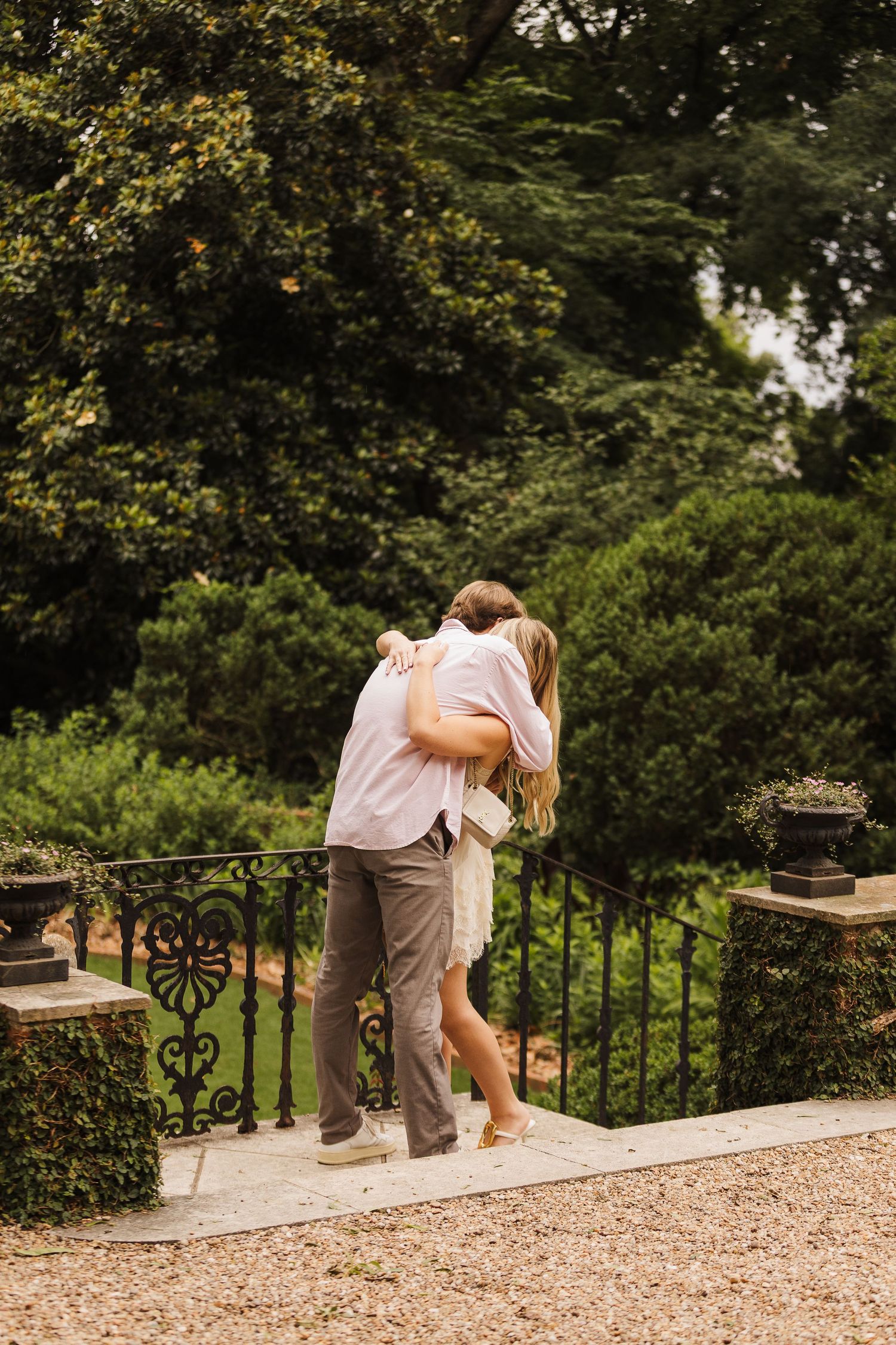 Couple embracing and kissing on a stone terrace surrounded by lush green gardens and ornate iron railings.