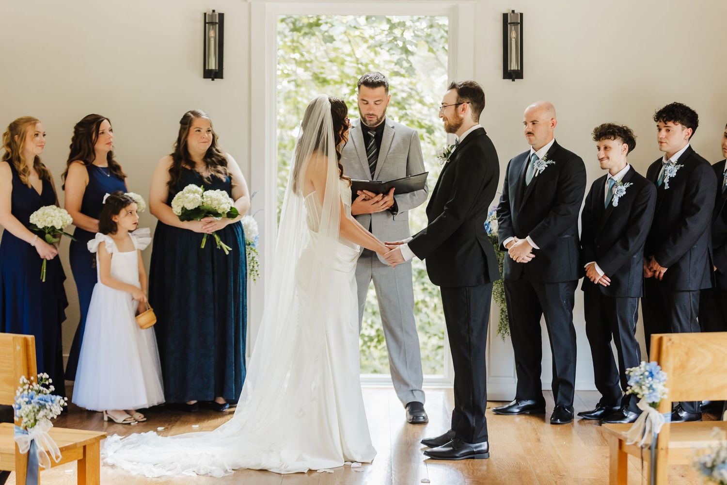 Bride and groom exchange vows at indoor wedding ceremony with bridesmaids in navy dresses and groomsmen in black suits.