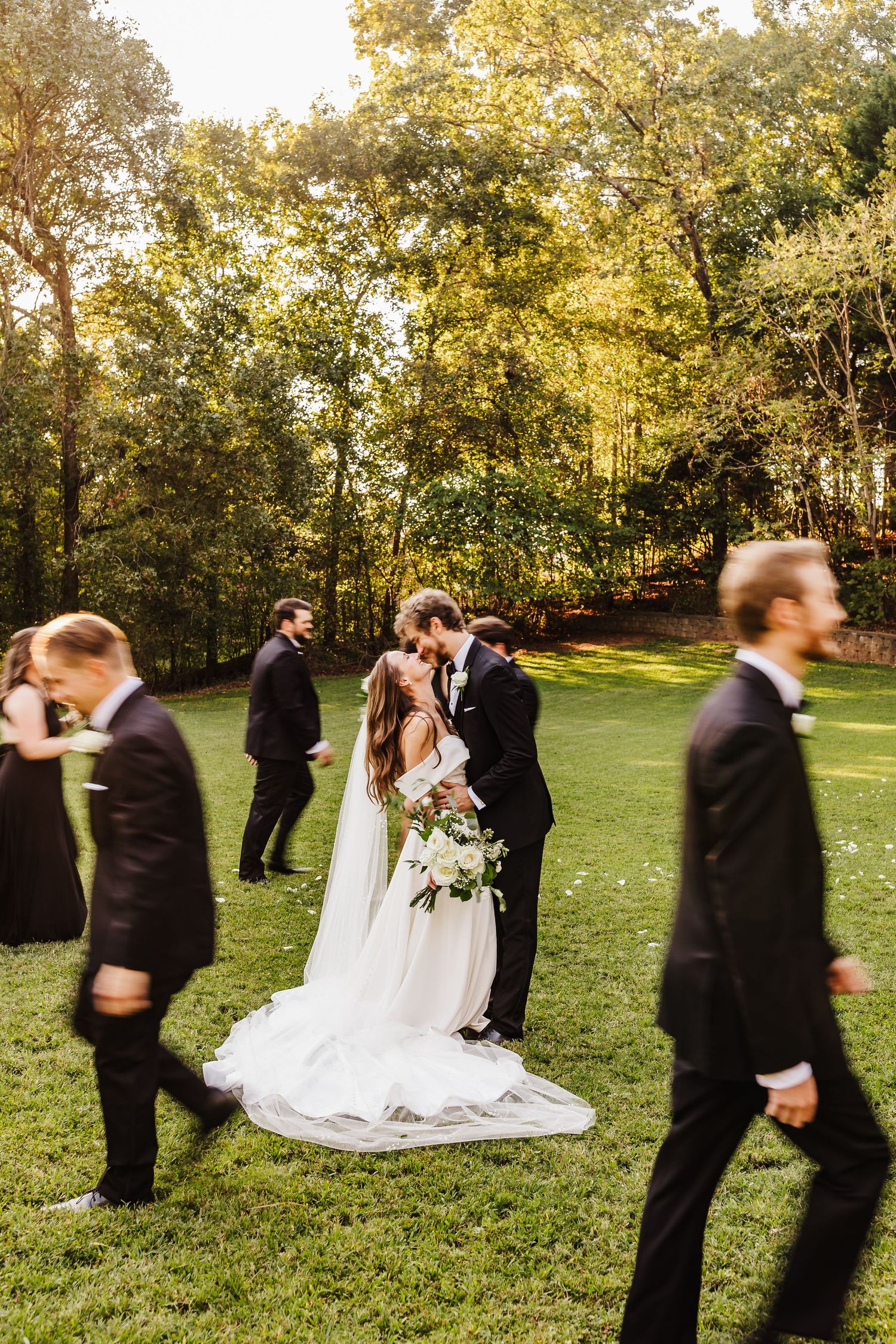 A wedding ceremony in a sunlit outdoor garden with bridesmaids and groomsmen in black suits.