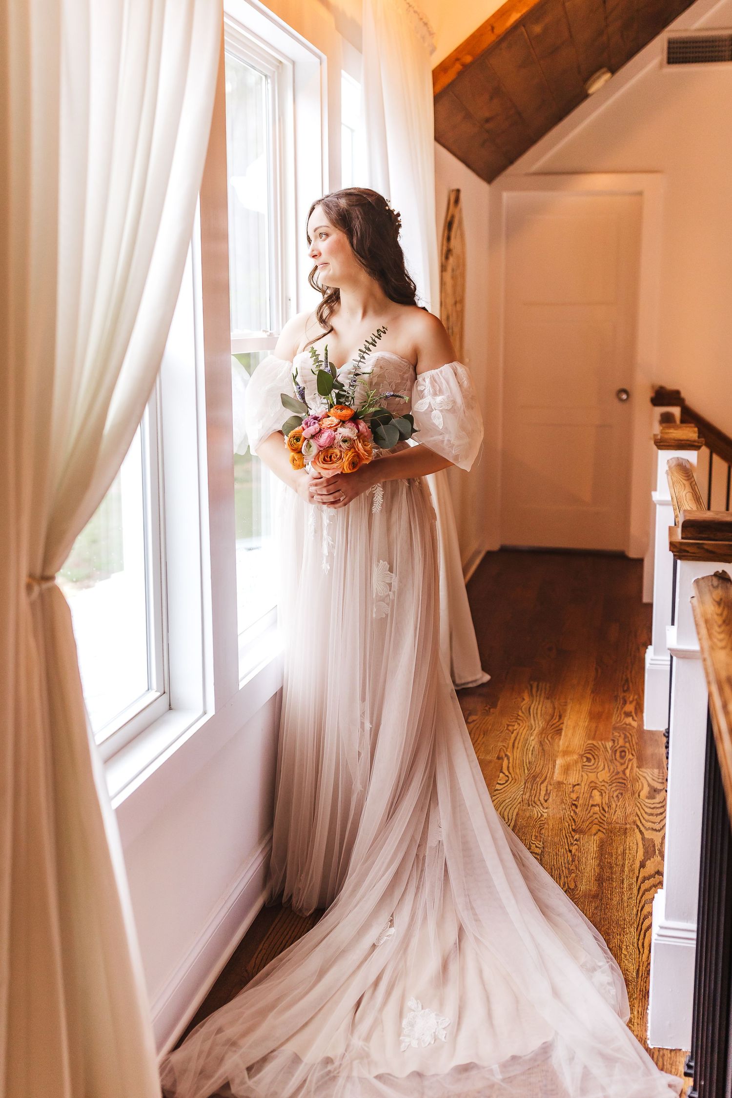 Flowing white wedding dress with train captured in a sunlit room with white curtains.