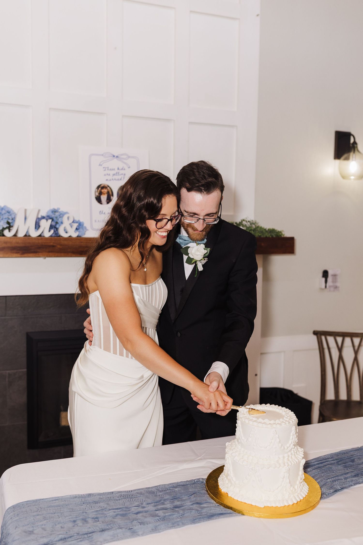 Bride and groom cutting white wedding cake together at reception, wearing glasses, with blue floral decor and fireplace backdrop.