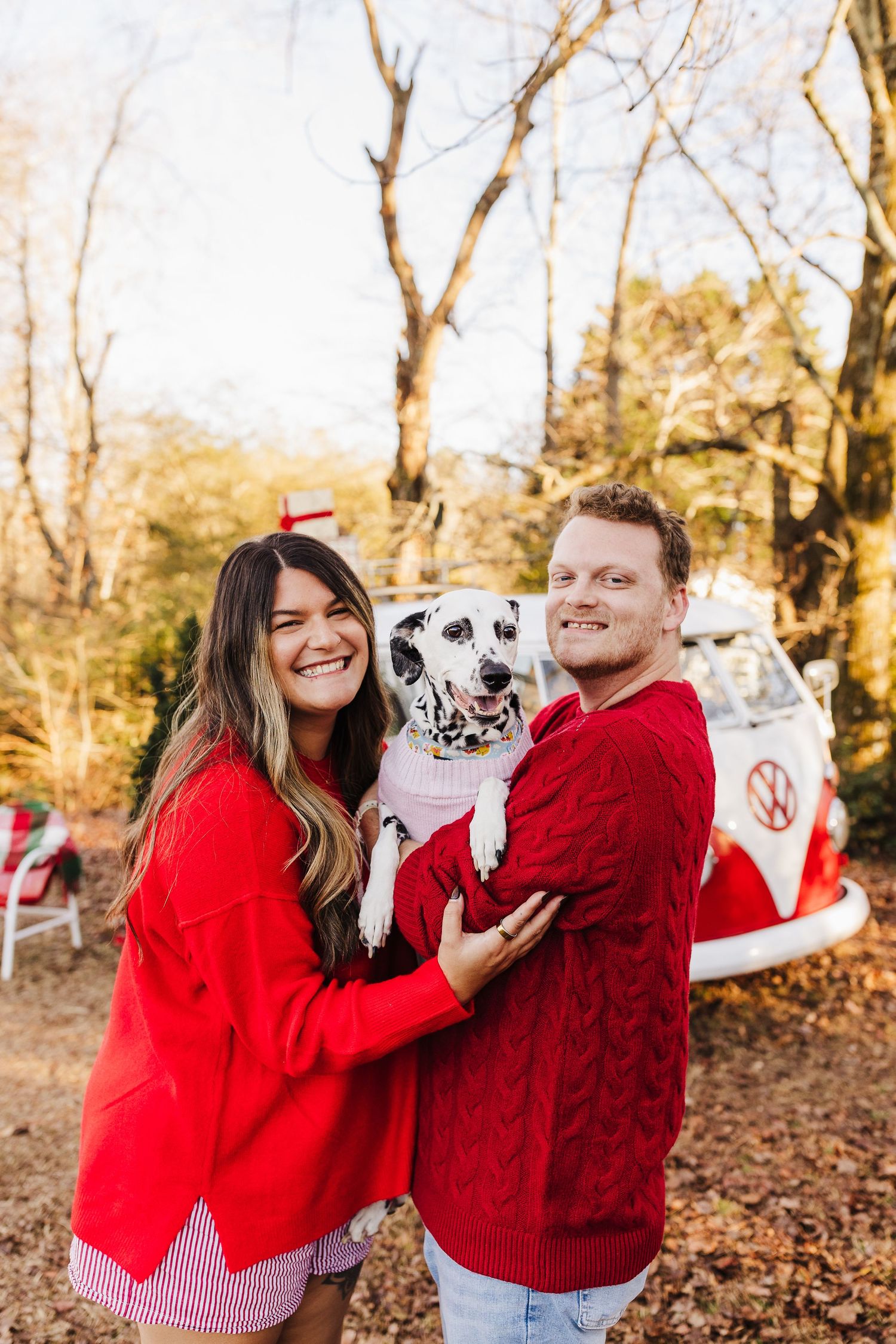 A smiling couple in matching red sweaters holds a Dalmatian dog outdoors near a vintage VW bus in autumn.